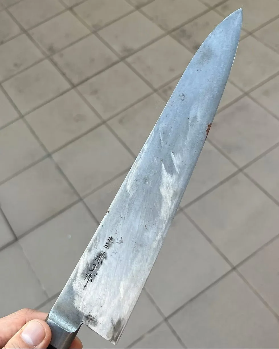 Een metalen koksmes met Japanse karakters erop, vastgehouden door een hand, met een betegelde vloer op de achtergrond.