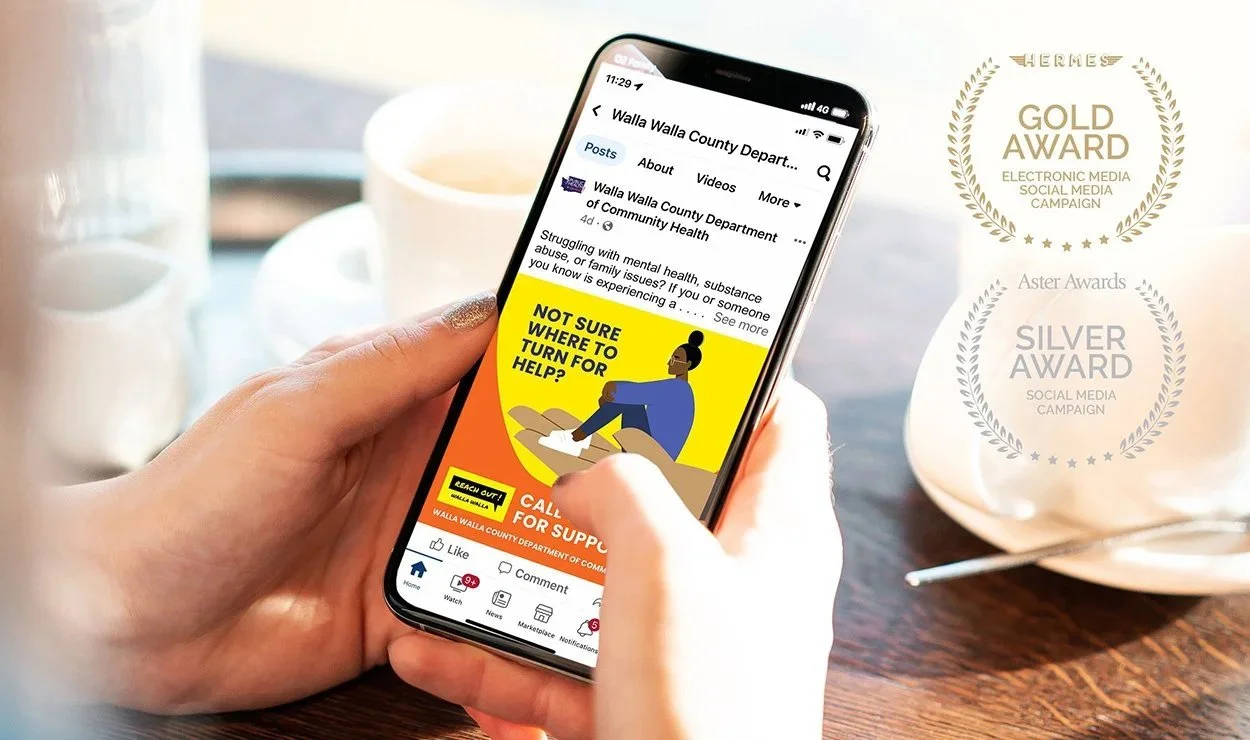 Person holding a smartphone displaying a Facebook post about mental health support with a cartoon of a woman sitting and a yellow background. In the background, there is a white mug and a glass on a wooden table.