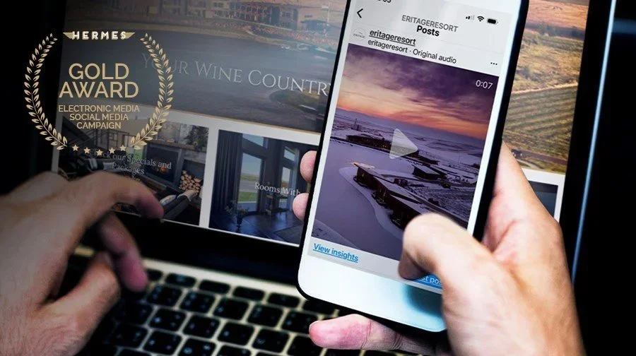 Person holding a smartphone displaying a sunset over snow-covered harbor, with a play button indicating a video, in front of a laptop showing a webpage with an award badge and images of a hotel or resort.