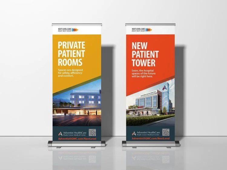 Two vertical banners in a display stand. The left banner is orange and blue and promotes private patient rooms, with an image of a hospital building at the bottom. The right banner is red and blue and announces a new patient tower, with a different h
