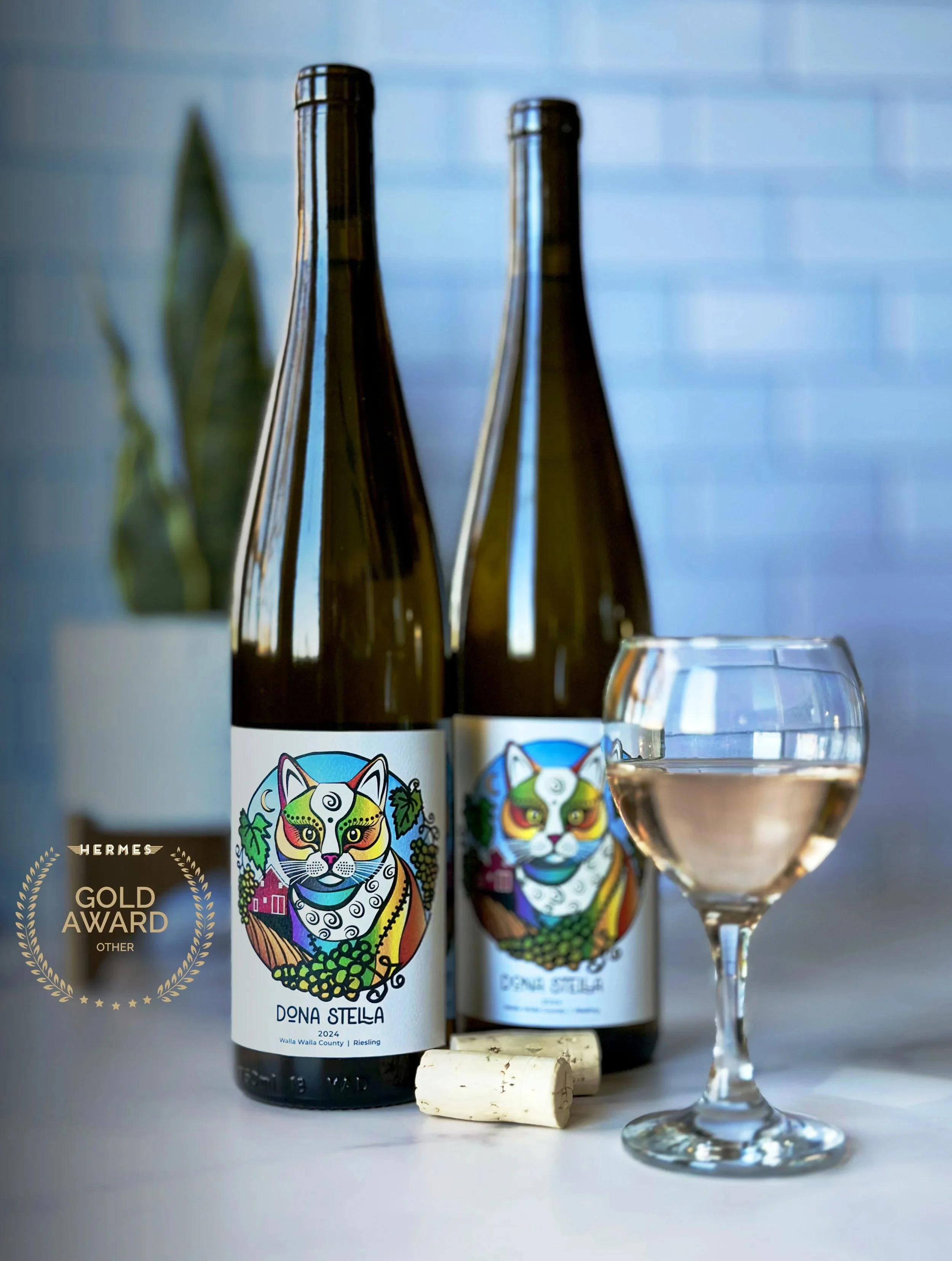 Two wine bottles with colorful, artistic labels featuring a feline face, positioned behind a glass of rosé wine, with a cork and additional cork in the foreground, and a blue-tiled wall in the background.