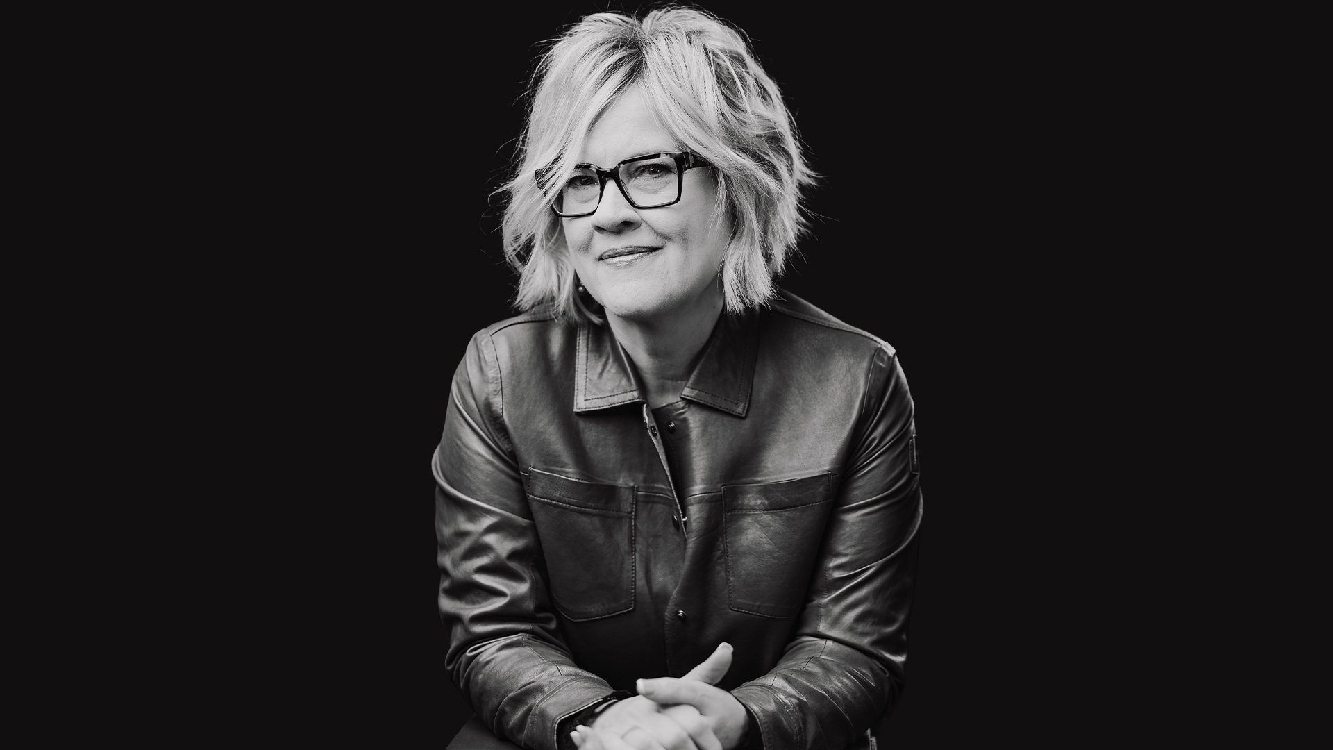 Black and white portrait of a woman with short, wavy blonde hair, wearing glasses and a leather jacket, sitting against a dark background.