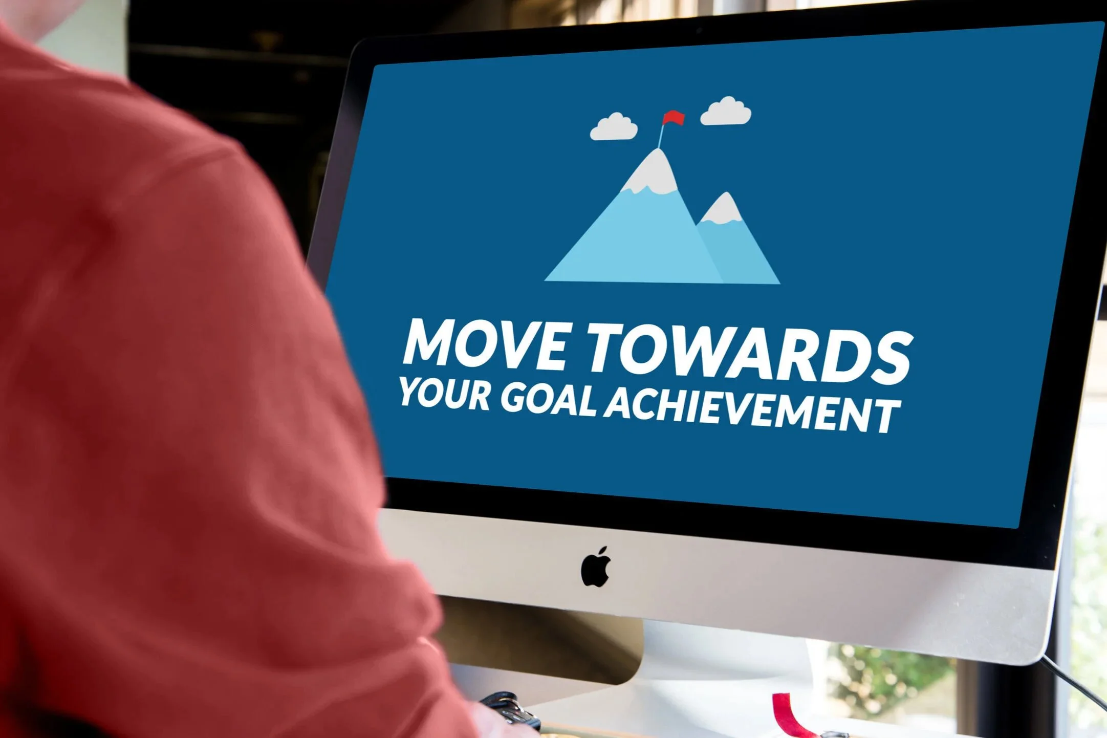 A person is holding an iMac computer displaying a graphic with mountains, clouds, and a red flag. The graphic includes the text 'Move Towards Your Goal Achievement'.