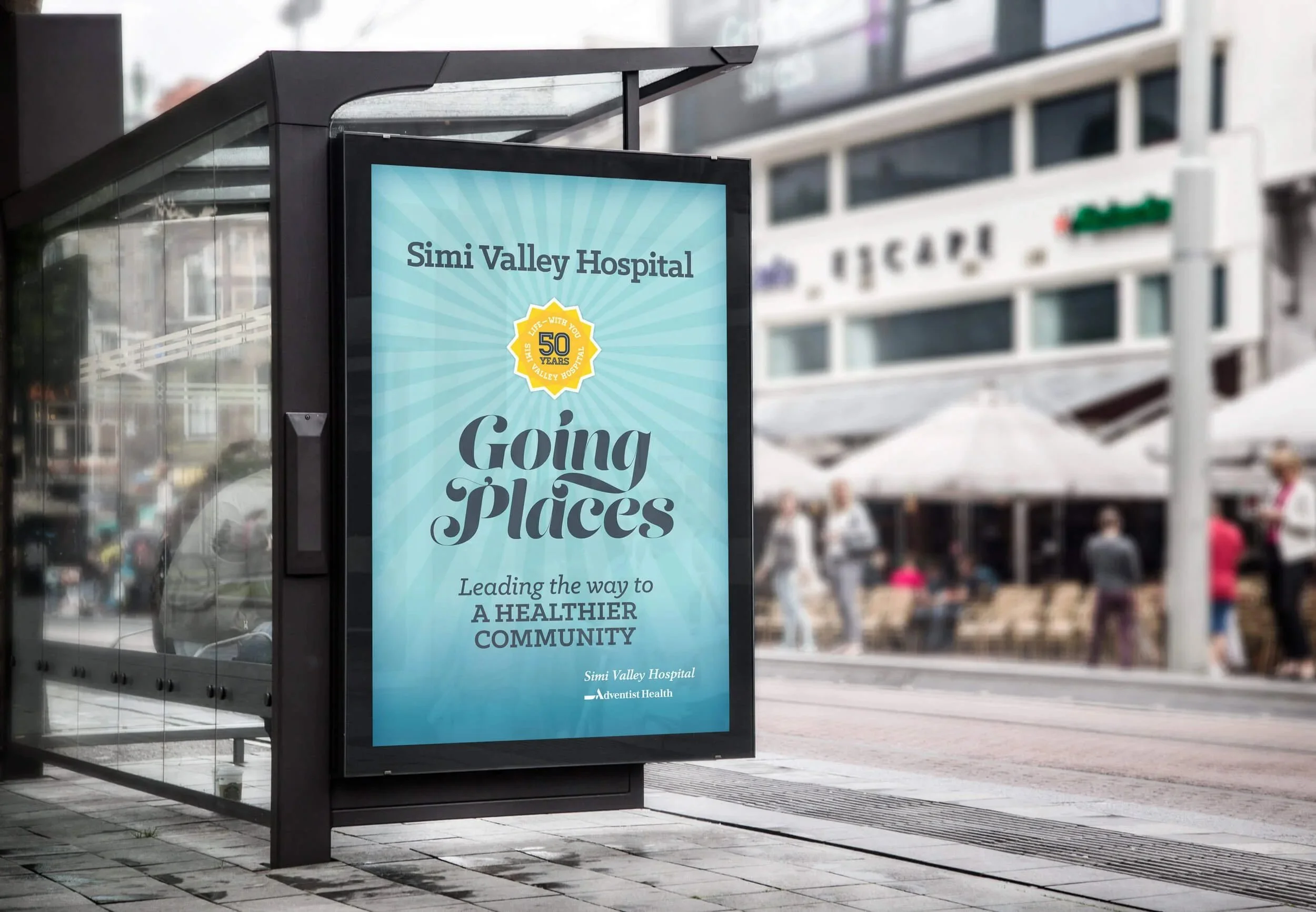 Bus stop shelter with an electronic display advertising Simi Valley Hospital's 50th anniversary and community health initiative, with buildings and pedestrians in the background.