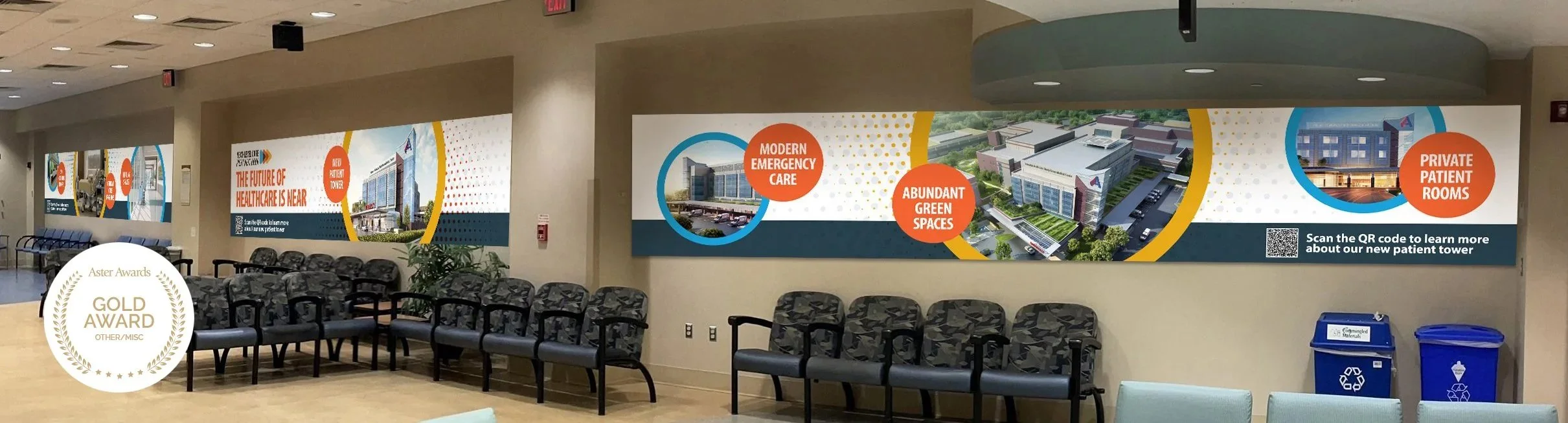 Hospital waiting area with chairs and large informational banners about healthcare facility features including emergency care, green spaces, and patient rooms.