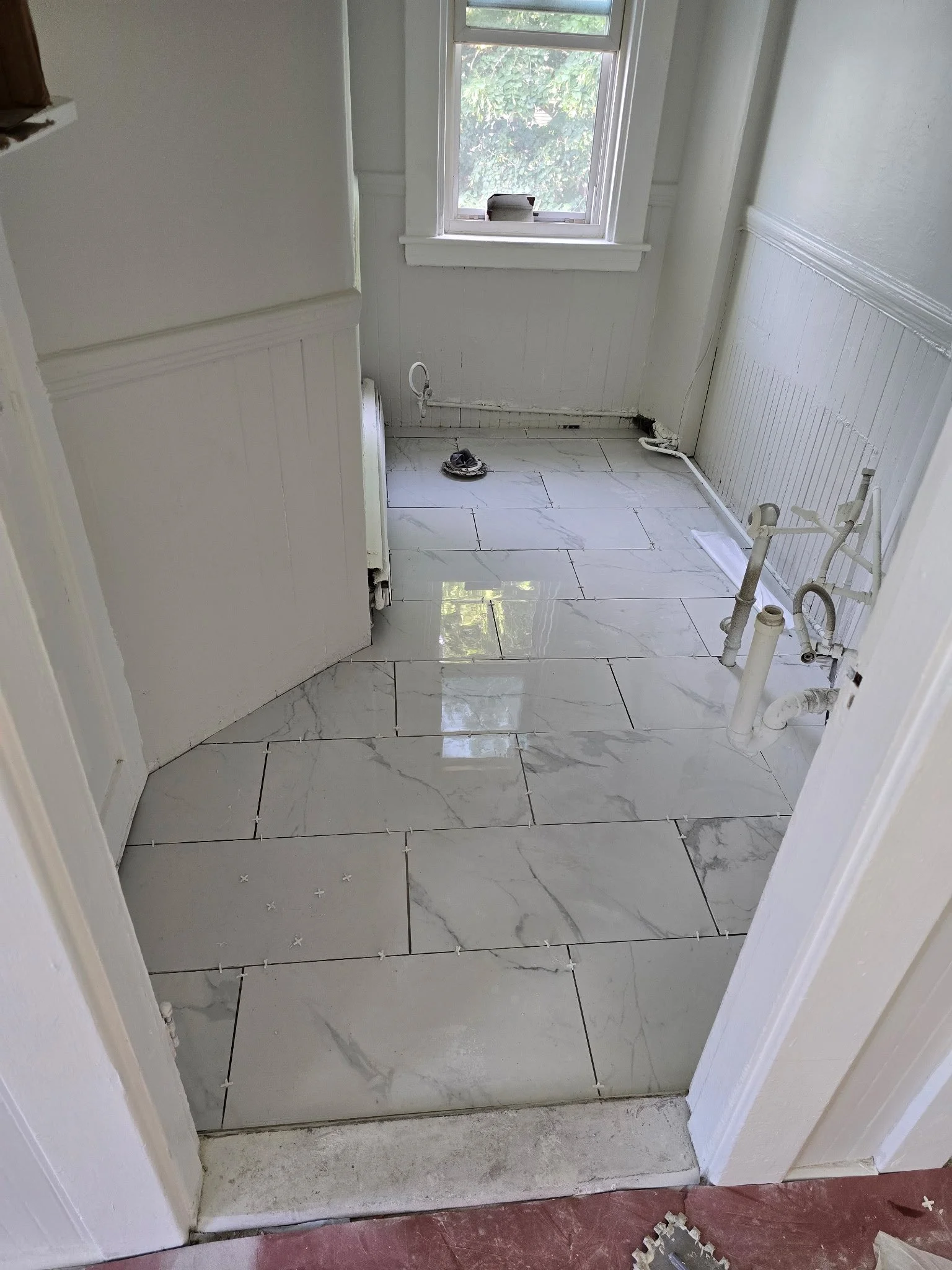 A small room with white walls and large white marble-patterned tiles on the floor. There is a window with a green leafy view outside. Construction materials and pipes are visible, indicating ongoing renovation or plumbing work.