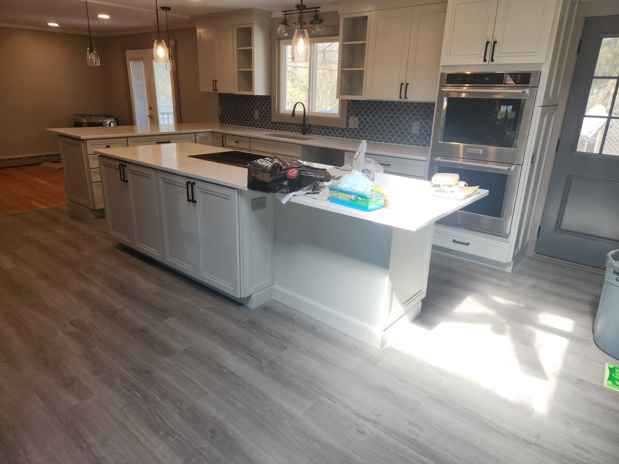 Modern kitchen with white cabinets, black hardware, a gray backsplash, and a large island with a white countertop. There are pendant lights hanging over the island and a window above the sink showing outdoor trees. Various items are scattered on the island, including a plastic bag and wipes.