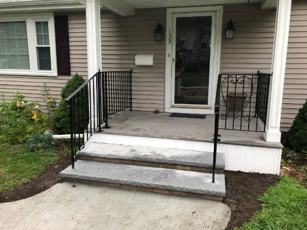 Front porch of a house with steps leading up to a door, black metal railings, potted plant, mailbox, and outdoor lighting fixtures.
