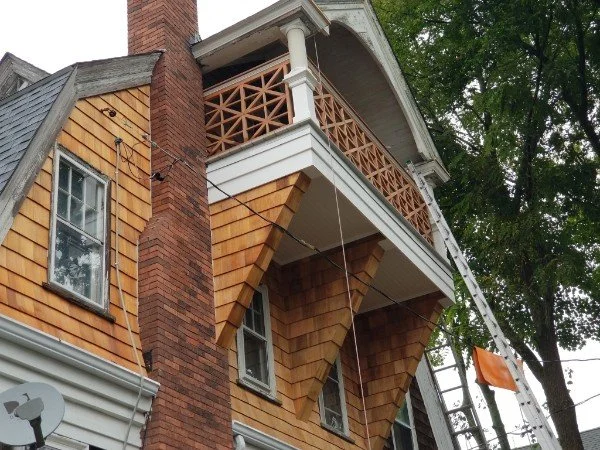 Close-up of a multi-story house with a brick chimney, wooden shingle siding, and a wraparound balcony with decorative wooden railing, surrounded by green trees.