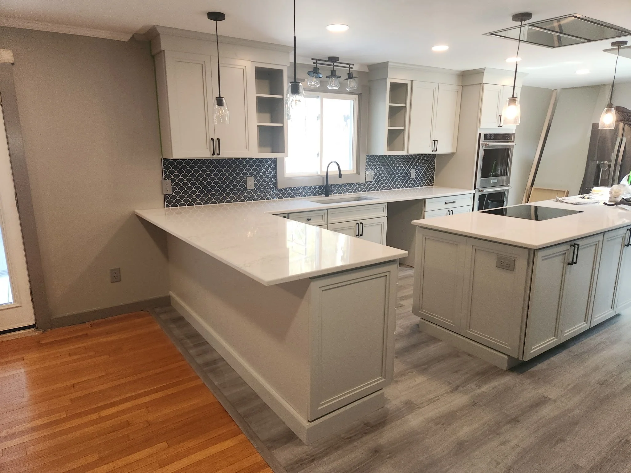 A modern kitchen with white cabinets, a black fish scale backsplash, a window with light coming through, and pendant lights hanging from the ceiling.