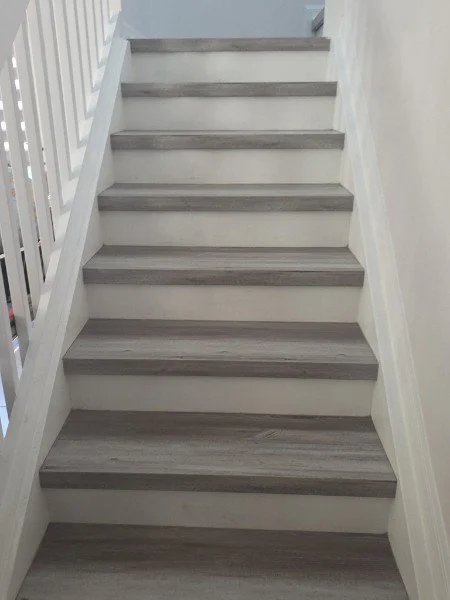 A straight staircase with gray wooden treads and white risers, bordered by white walls and a white railing on the left side.