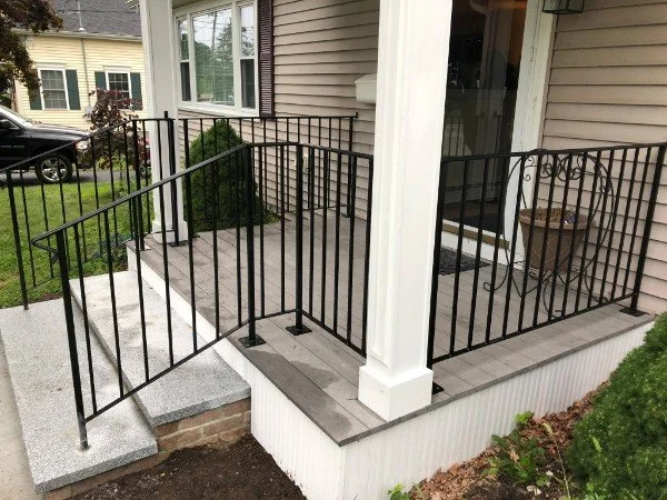 Front porch with black metal railing, concrete steps, and a white support column in front of a house with beige siding.