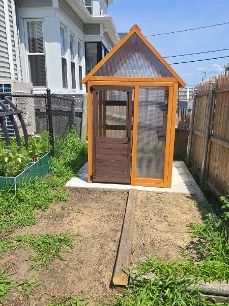 A small wooden greenhouse with a sloped roof, glass sides, and a sliding door, situated on a concrete pad in a backyard with a garden on the left and a wooden fence on the right.