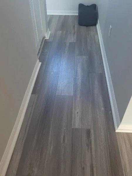 A narrow hallway with gray wood flooring and light-colored walls, with a black bag or cushion placed on the floor at the end of the hallway.