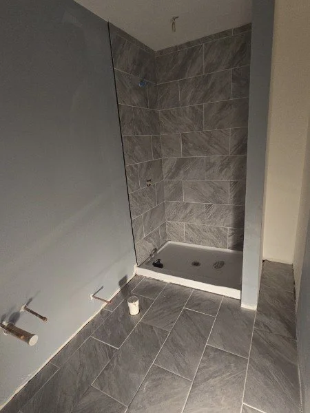 Empty shower stall with gray tiled walls and floor, and a partially installed wall on the left, with plumbing fixtures nearby.