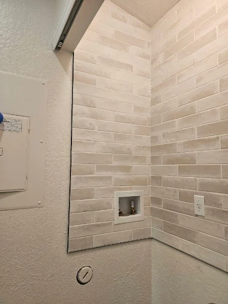 Corner of a room with beige brick-patterned tile wall, an electrical outlet, and a small built-in niche with plumbing hookups.