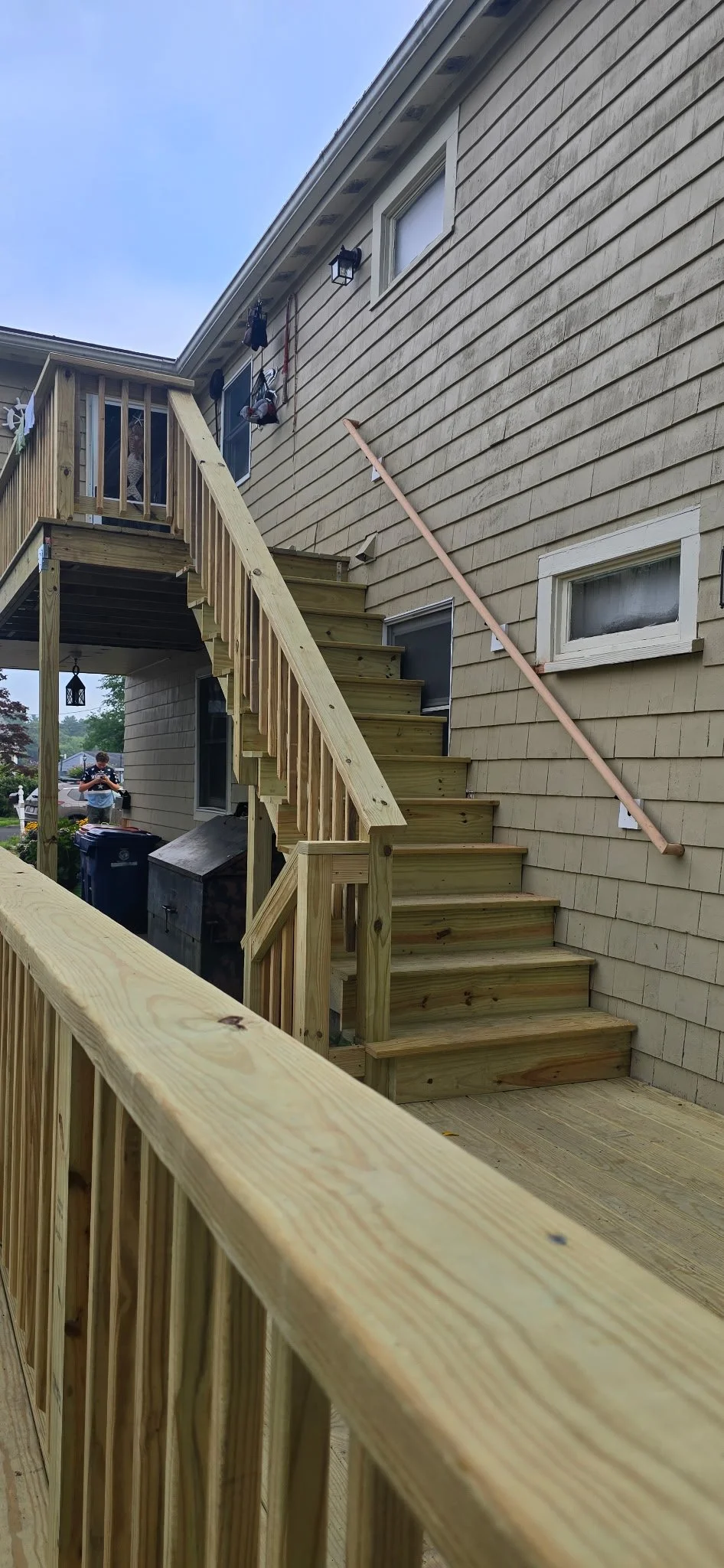 Newly built wooden staircase with railing leading from a deck to the yard of a house with beige siding, windows, and outdoor lights. A person is visible in the background near trash cans.