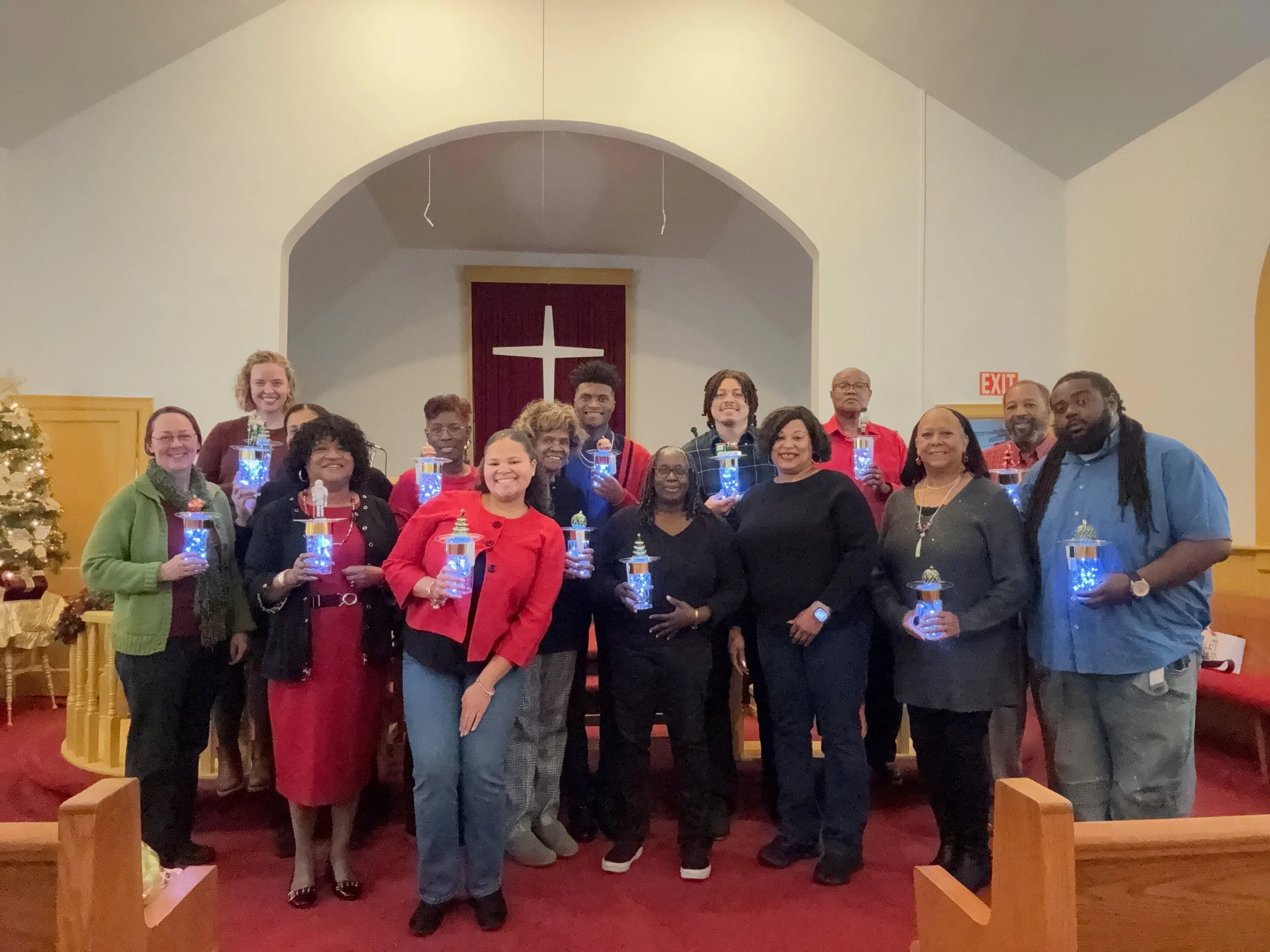 A group of 18 diverse people standing together inside a church, holding small lanterns with blue lights, smiling for a group photo during a Christmas celebration. In the background, a cross is visible on the wall, and there is a Christmas tree on the