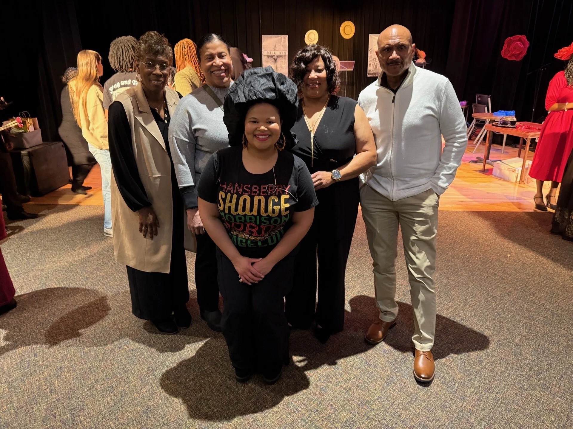 Group of six people, five women and one man, standing together in a dark room, smiling for the camera. The woman in front is wearing a black shirt with colorful text and a large black hat. The man is wearing a white jacket and tan shoes. The women ar