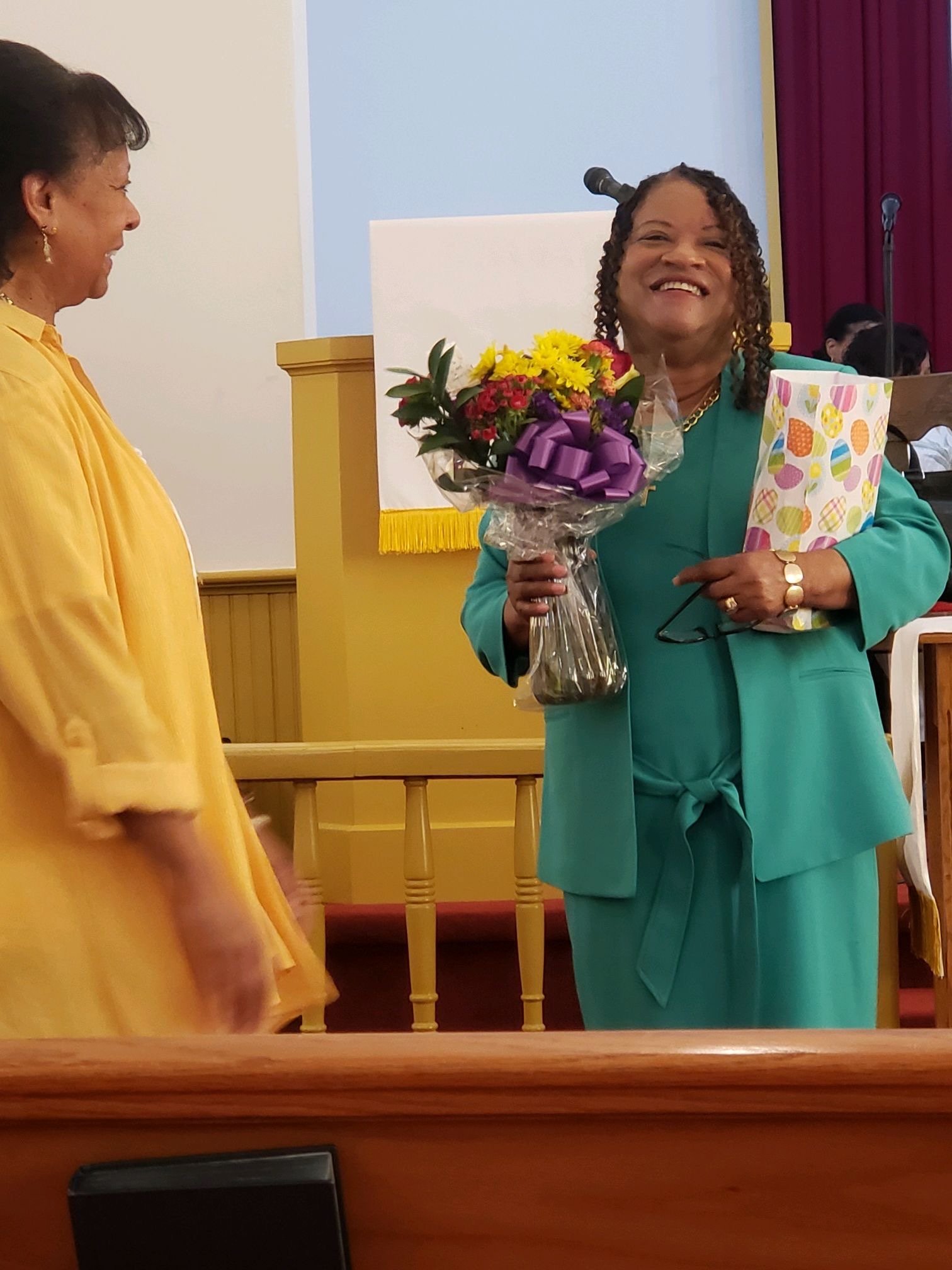 A woman with curly hair smiling and holding a bouquet of flowers and a wrapped gift while standing at a church podium. Another woman in yellow is partially visible on the left. The setting appears to be a celebration or special event.