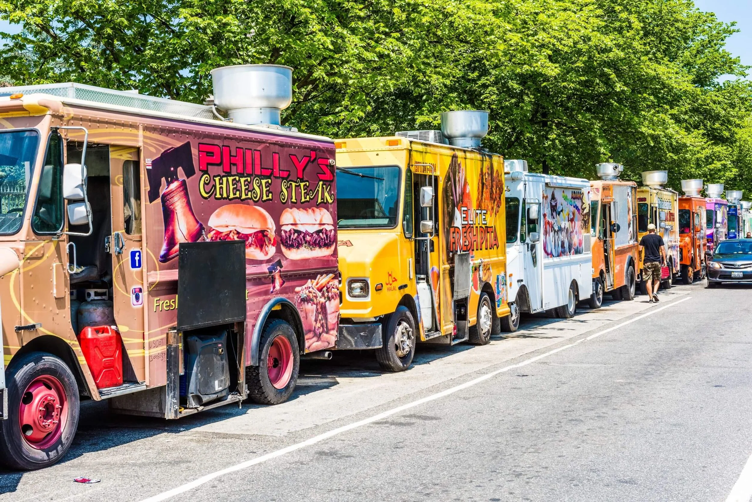 Line of colorful food trucks parked along a street with green trees in the background, including a truck advertising Philly's cheese steak.