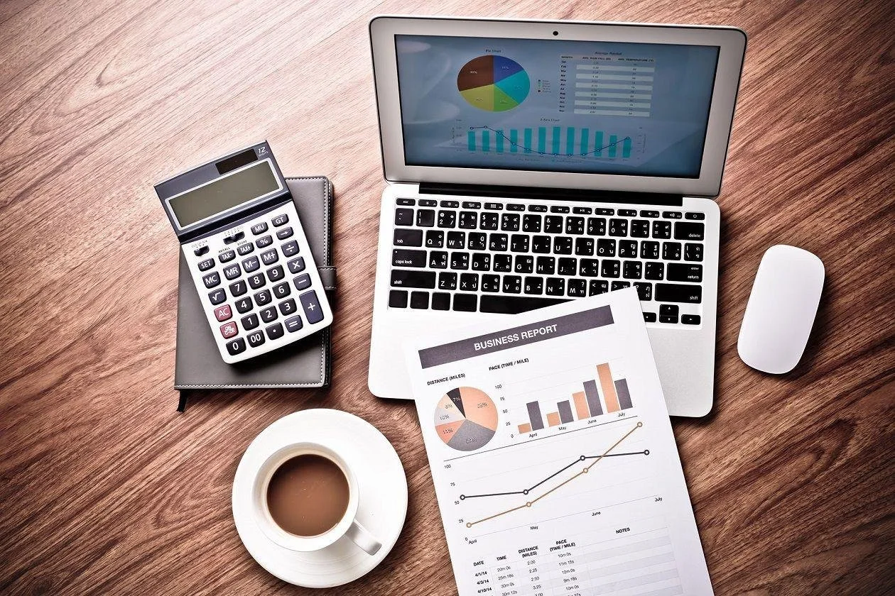 Work desk with a laptop showing graphs, a calculator on a notepad, a cup of coffee, a business report with charts, and a wireless mouse on a wooden surface.