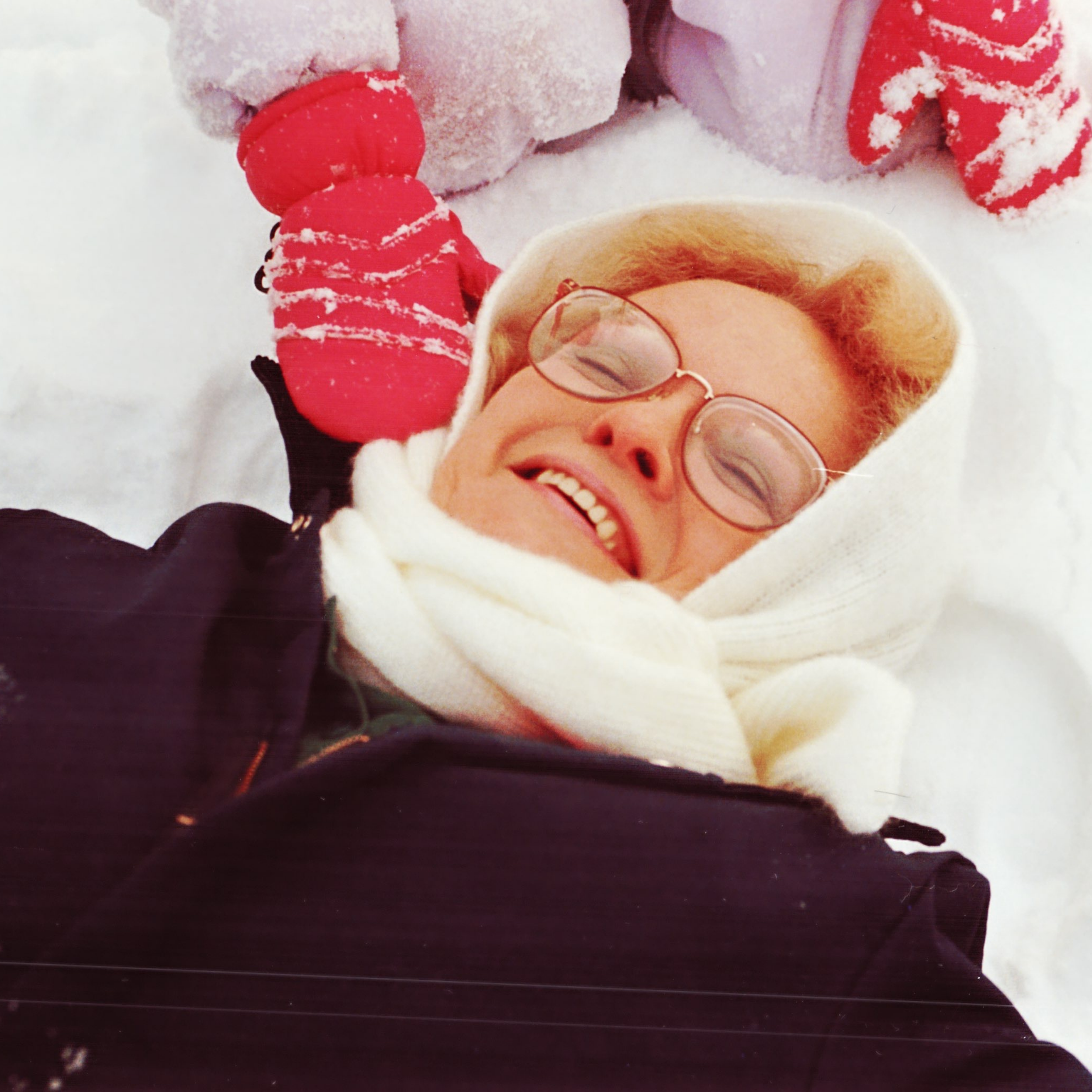 Smiling woman, bundled up, playing in snow while grandchild with red mitten pats her.