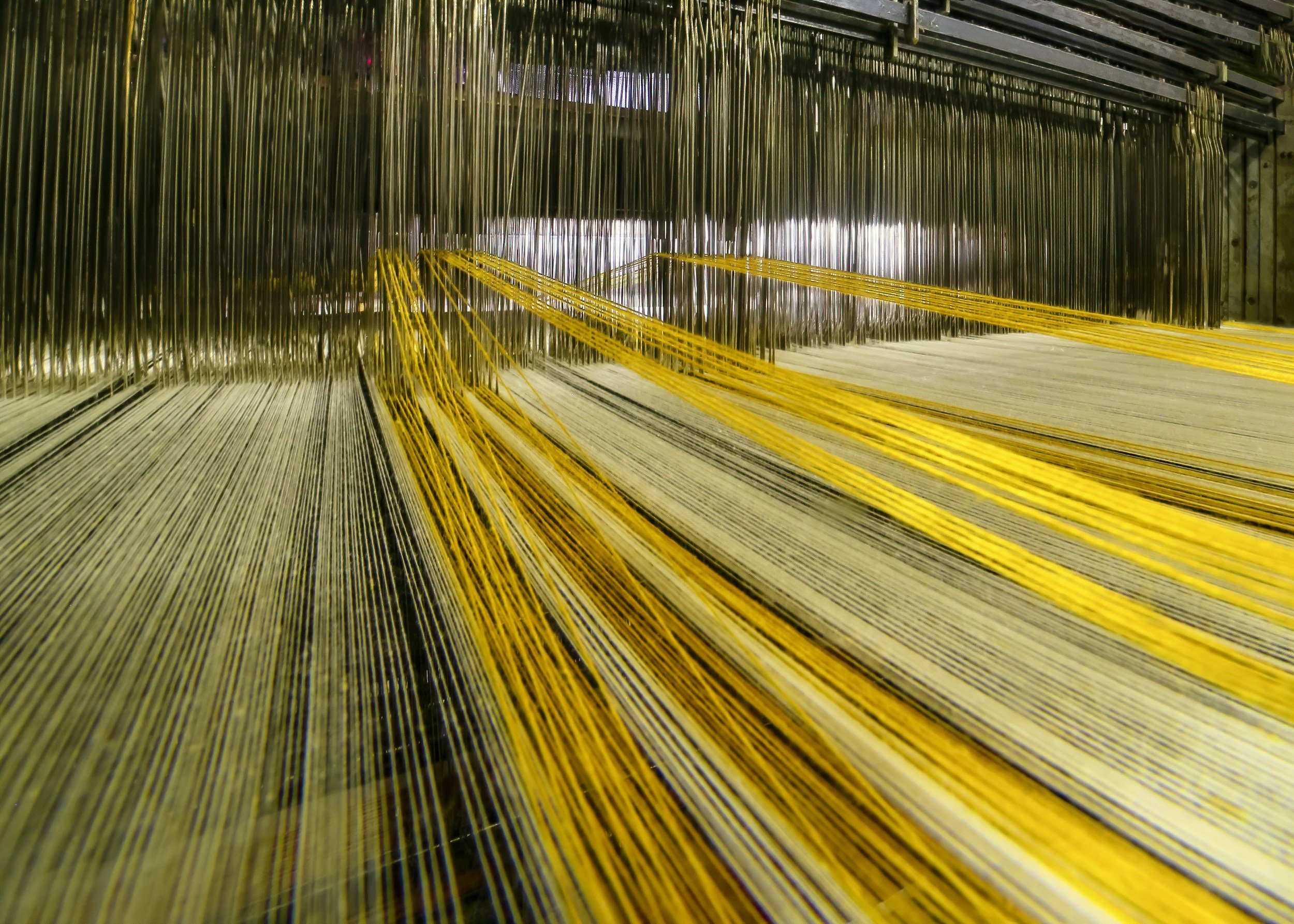 A weaving loom with yellow threads woven into fabric, surrounded by vertical metal rods and a wooden frame.