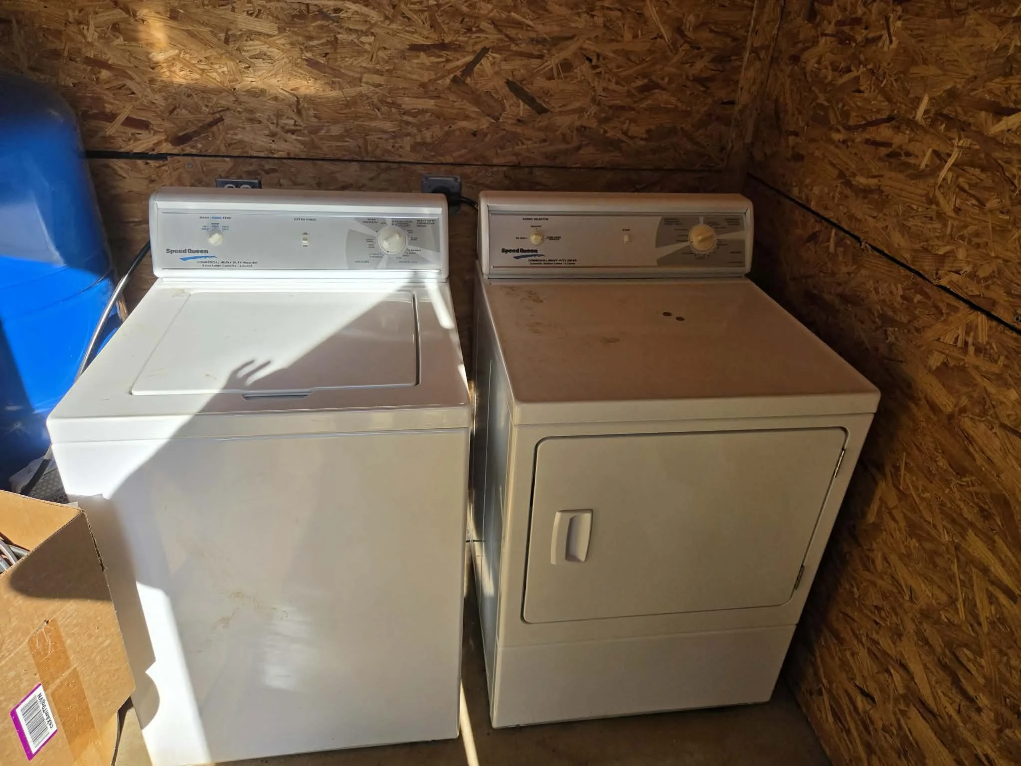 A white washing machine and beige dryer are placed side by side against a plywood wall in a laundry area. The washing machine lid is closed. There is a blue water jug on the left and a cardboard box on the bottom left corner of the image.