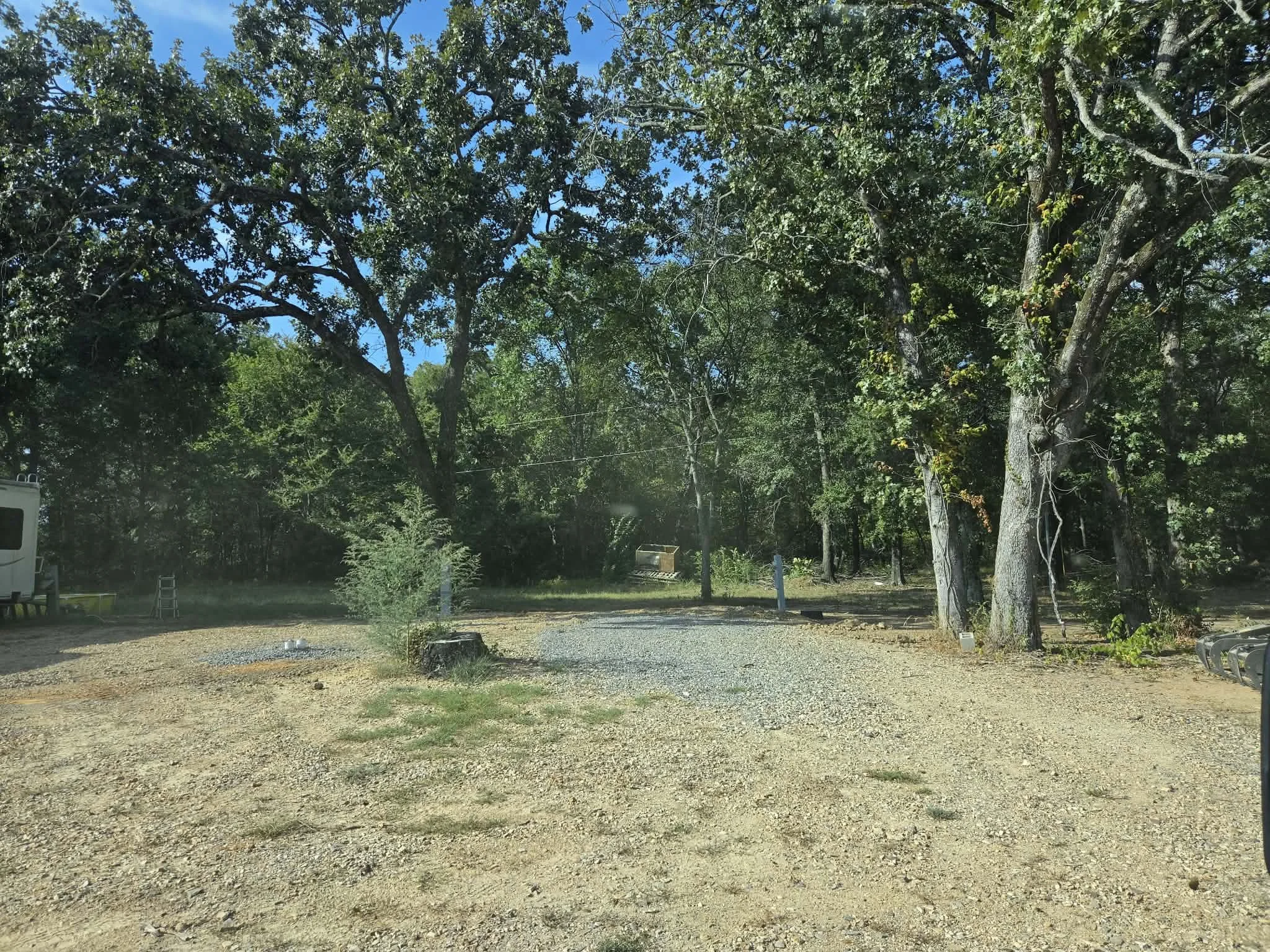 A gravel parking area surrounded by green trees and blue sky, with some outdoor furniture and a camper van in the background.