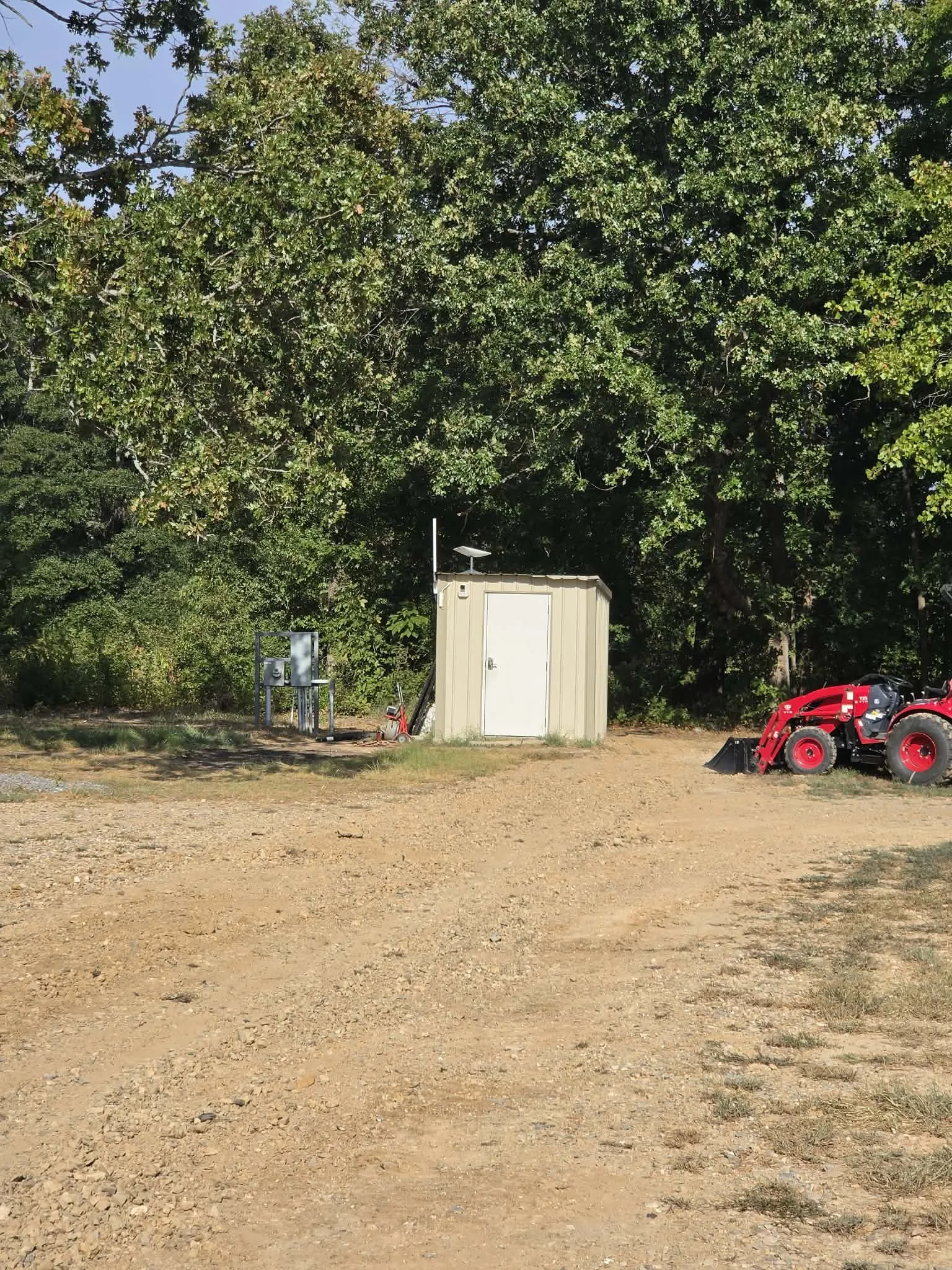 A dirt open space with some patches of grass, a small beige utility building, a red compact excavator, electrical boxes, and a background of large trees with green foliage under a clear blue sky.