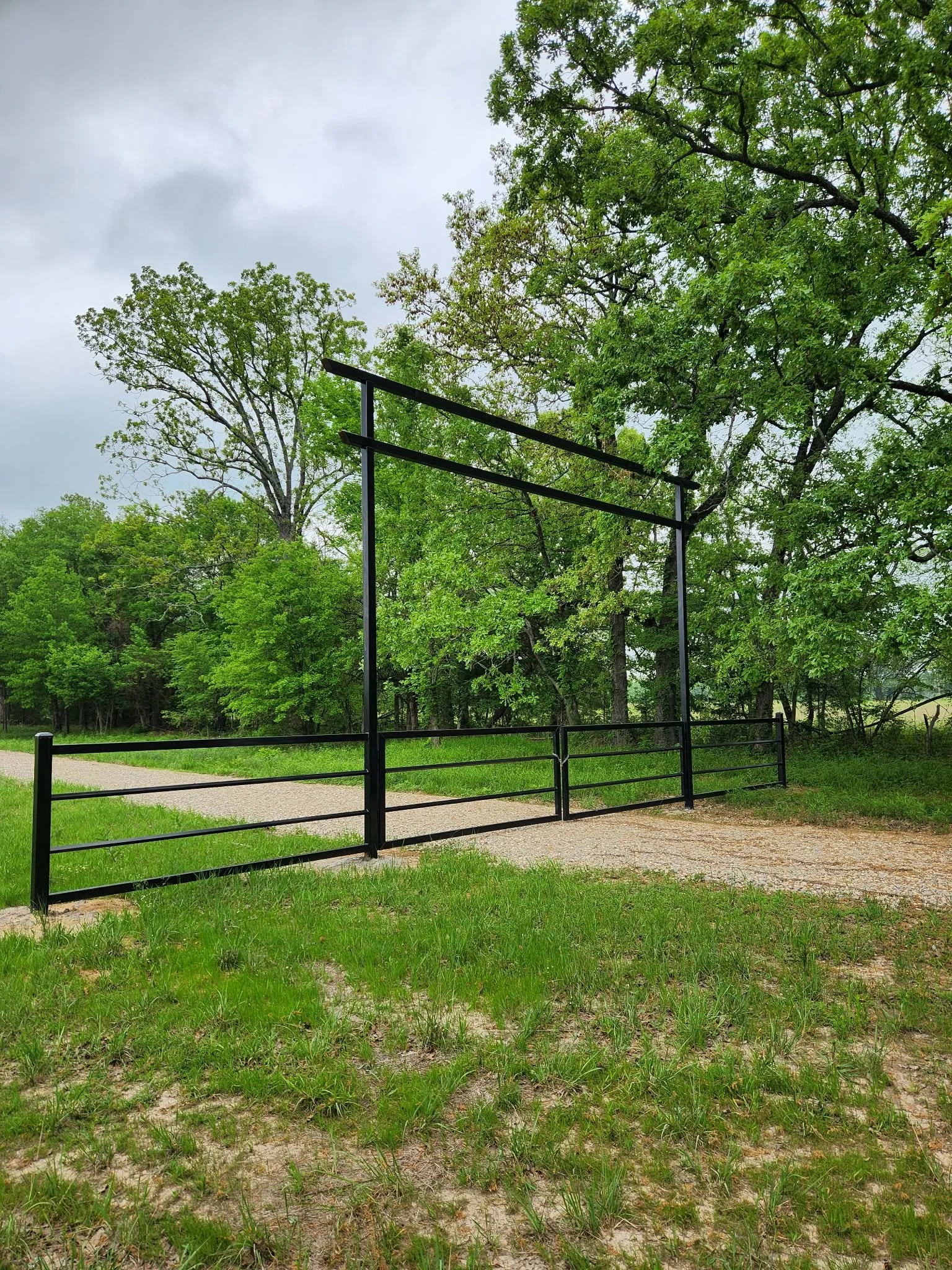 Empty outdoor basketball court with a black metal fence, surrounded by green trees and grass, under an overcast sky.