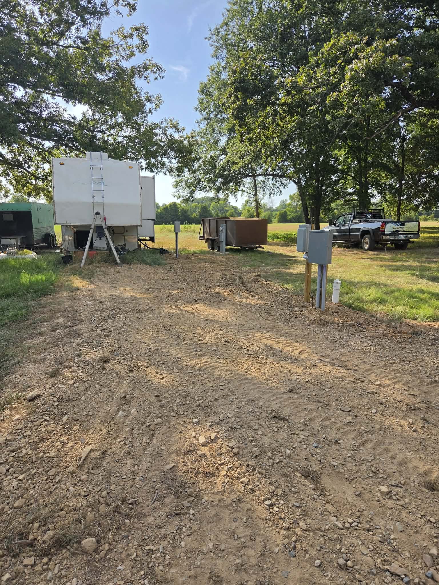 A rural scene with a dirt road, utility boxes, trailers, and a pickup truck parked under large trees on a sunny day.