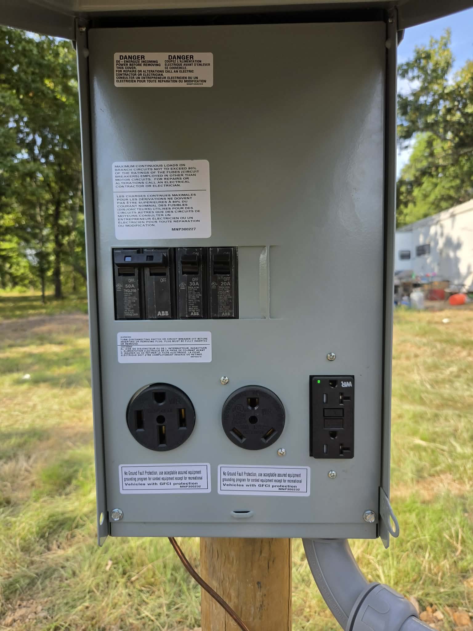 An outdoor electrical power station mounted on a wooden pole, with circuit breakers, power outlets, warning labels, and control switches, set against a background of trees and a partly cloudy sky.