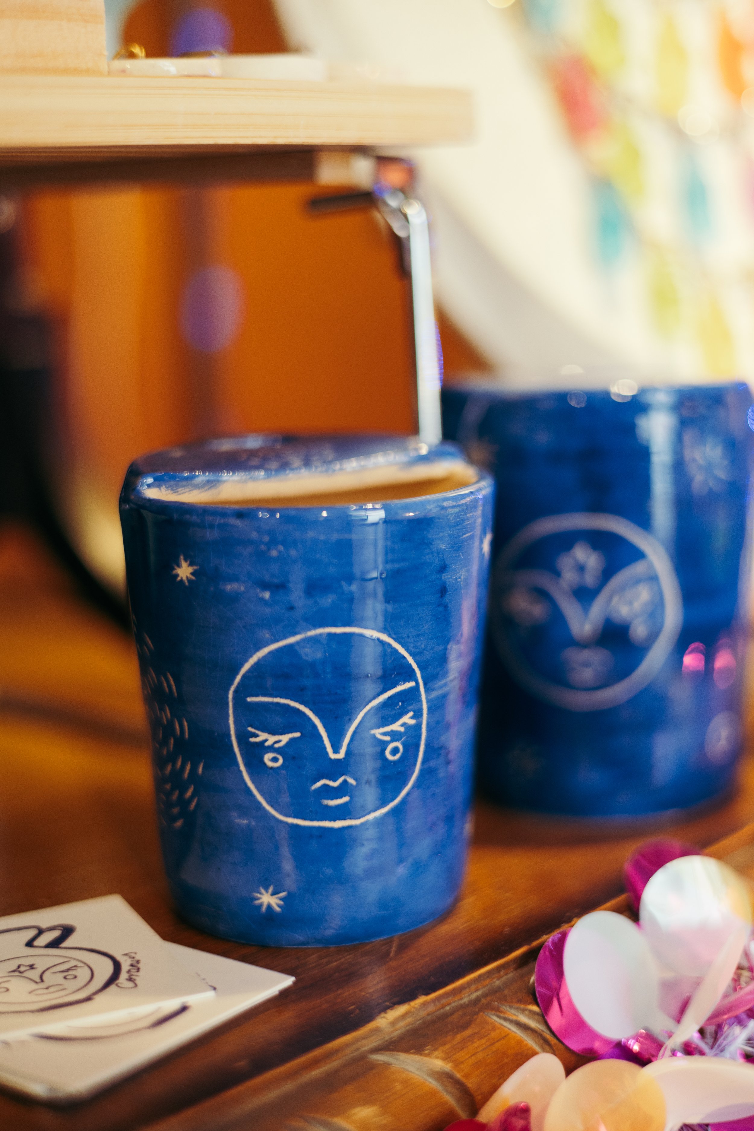 A blue mug with a white, abstract face design and star-like accents on a wooden surface, with colorful small decorative objects nearby.