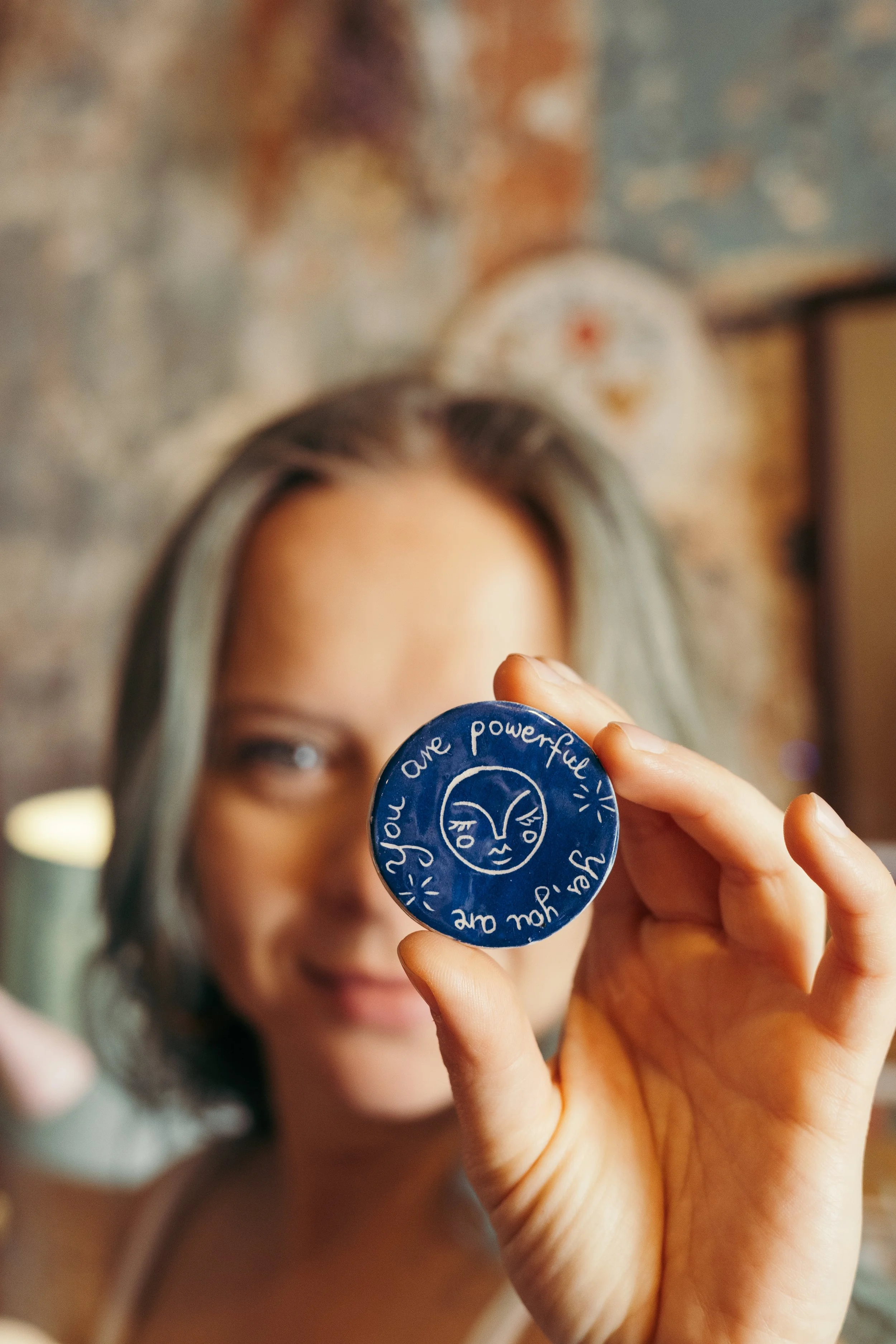 A person with glasses holds a blue circular sticker with handwritten words and a smiley face towards the camera, with their face blurred in the background.