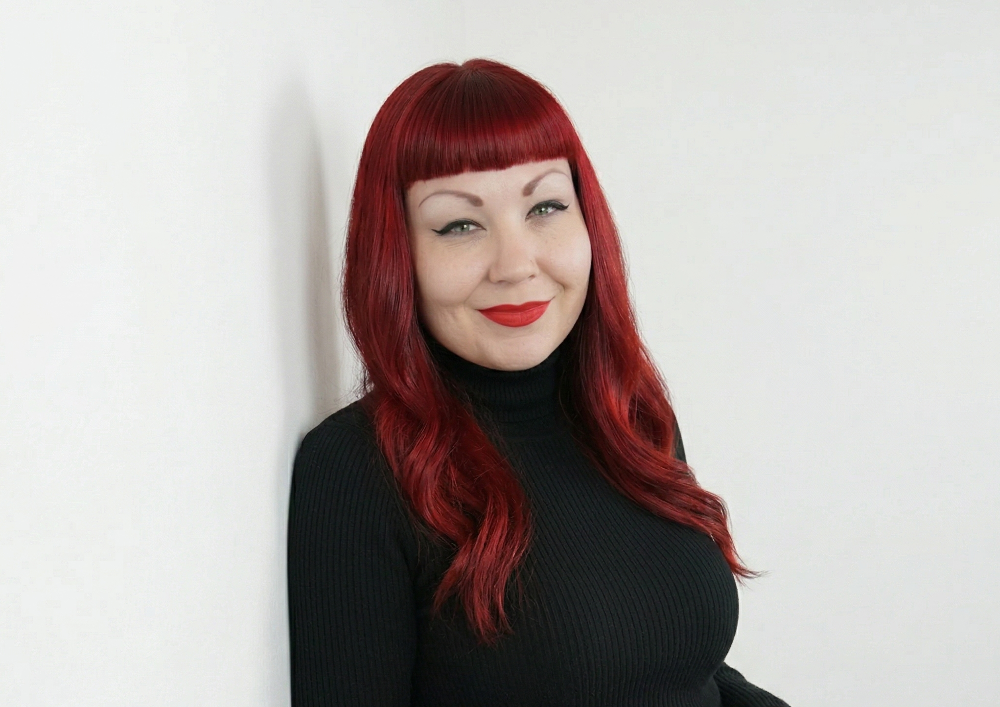 A woman with red hair and bangs smiling while wearing a black turtleneck against a white background.