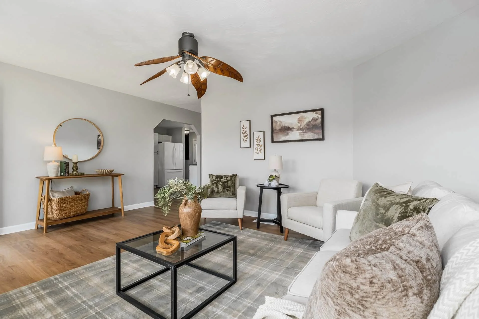 Living room with white sofas, a black coffee table with a decorative wooden sculpture, a potted plant, rug, wall art, a side table with a lamp, and a ceiling fan with wooden blades.