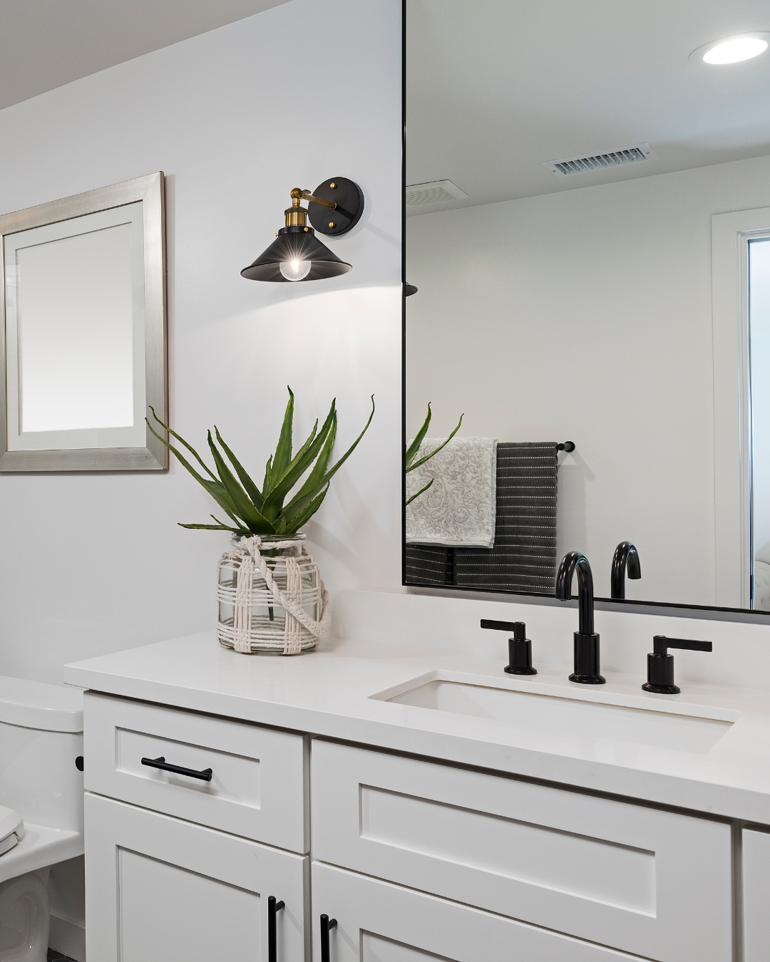 A modern bathroom with a white vanity, black faucet and hardware, a large mirror, a potted aloe vera plant, a towel rack with black and white towels, and a wall light fixture.