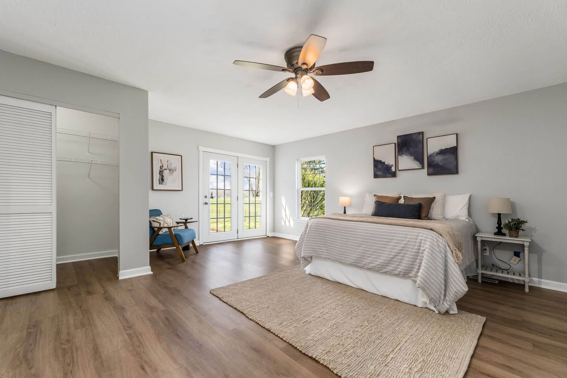 A bedroom with a white bed, side table and lamps, wooden floors, window and glass door, ceiling fan, and wall art.