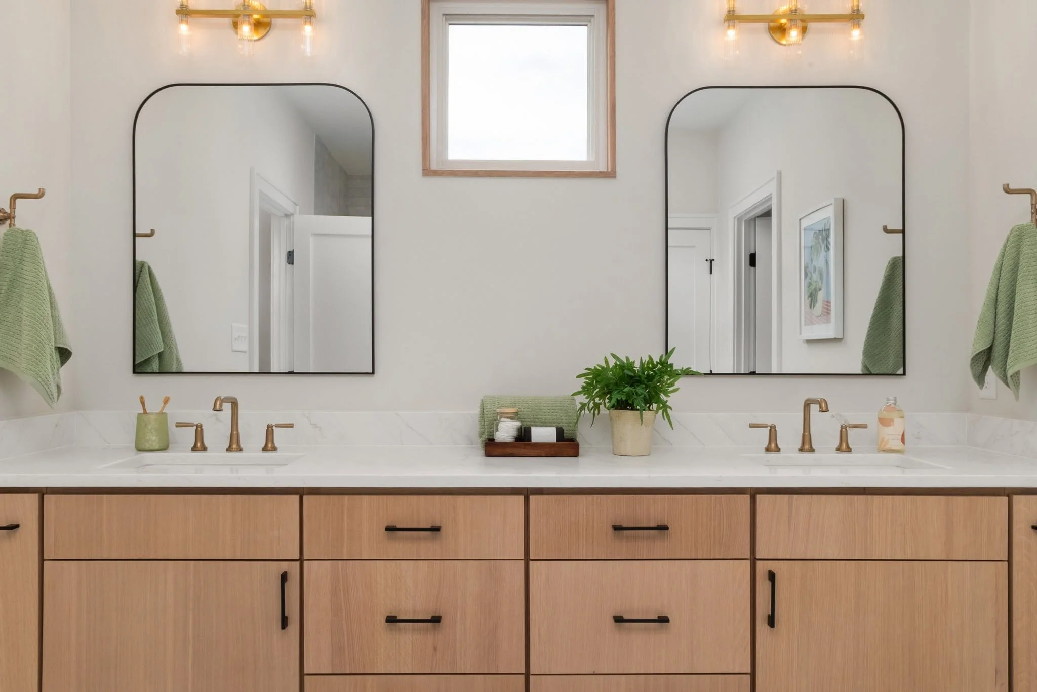 Double vanity bathroom with two mirrors, gold fixtures, a potted plant, green towels, and natural light from a window.