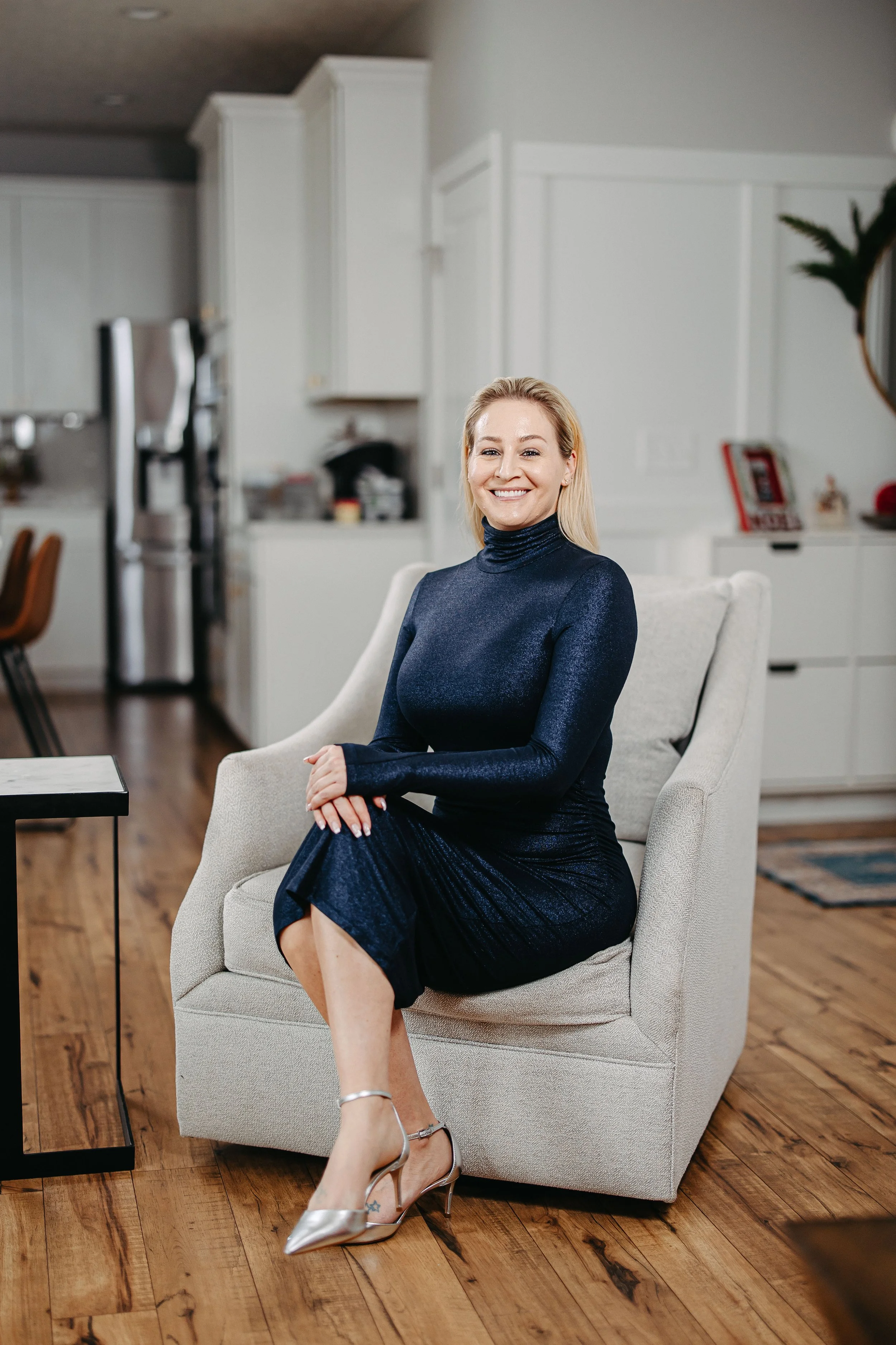 A smiling woman with blonde hair and wearing a sparkly dark blue dress is sitting on a beige armchair in a bright, modern living room with hardwood floors, a white kitchen in the background, and silver high heels.
