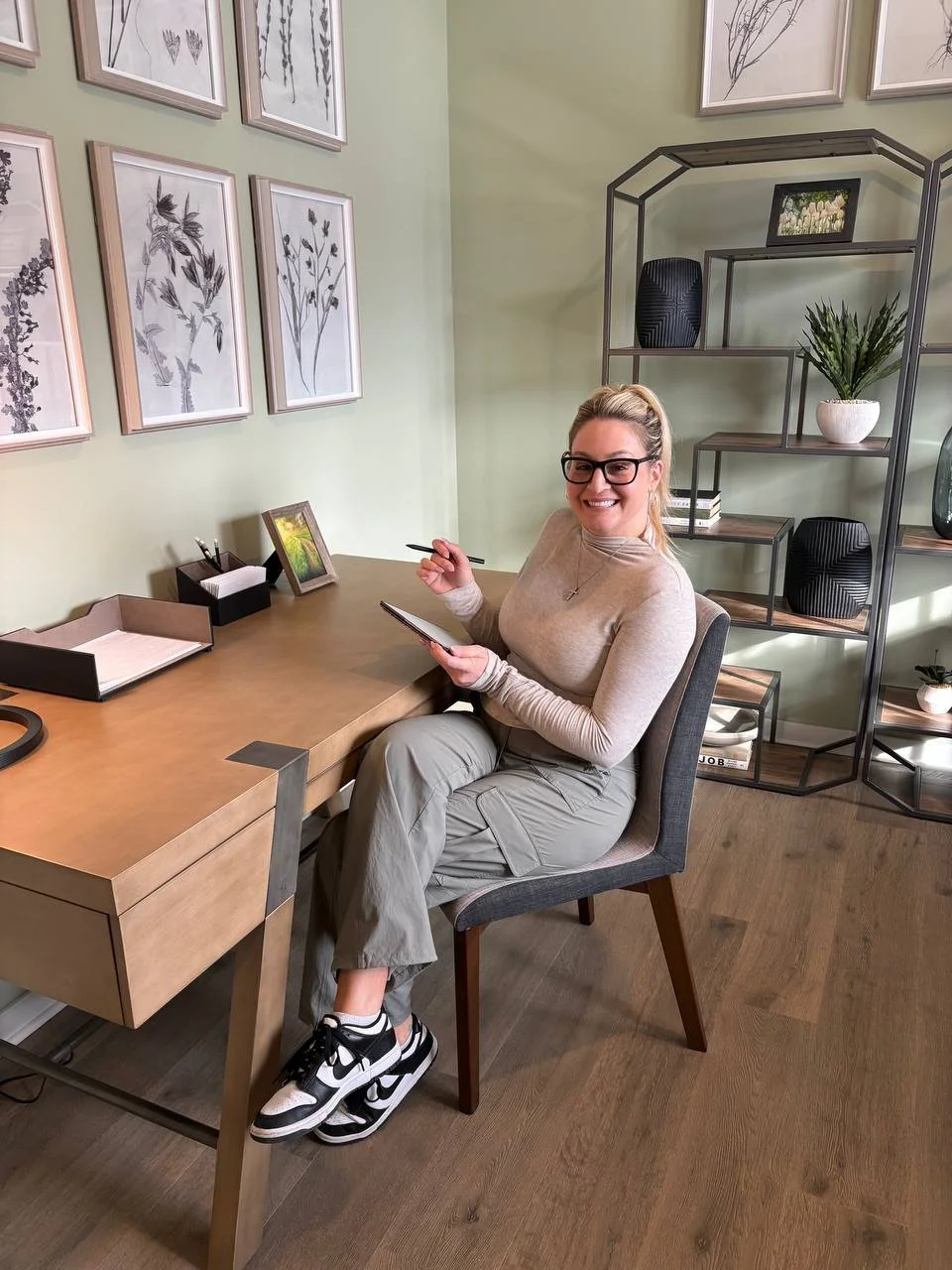 A woman with glasses seated at a desk in a modern office, smiling and holding a notebook and pen. The desk has a small picture frame, a black organizer, and a folder. Behind her, a green wall has framed botanical artwork, and a black metal shelf with decorative items such as a plant, a black vase, and framed photos.