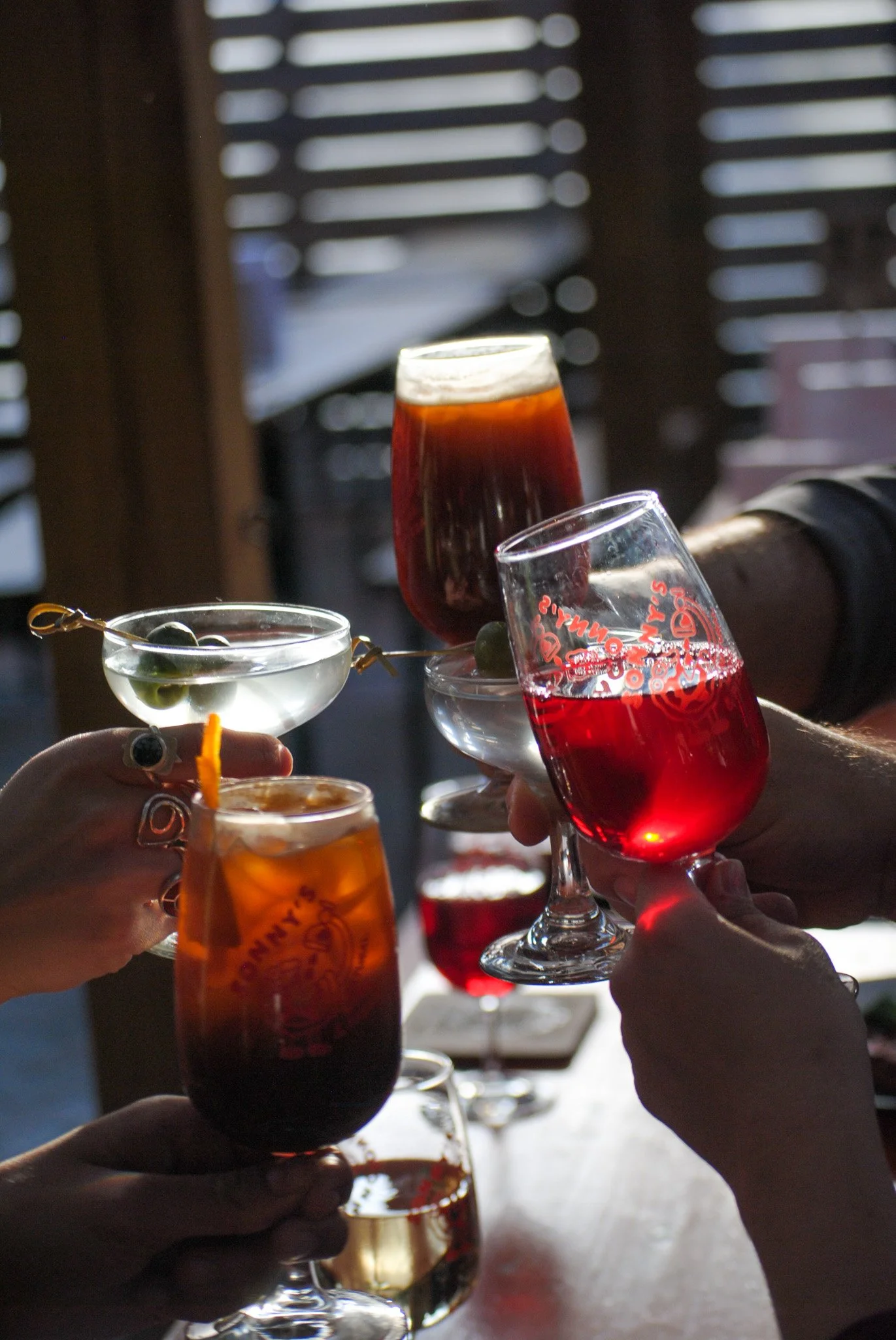 Several people raising glasses filled with colorful drinks in a toast inside a dimly lit room with wooden slats on the window in the background.