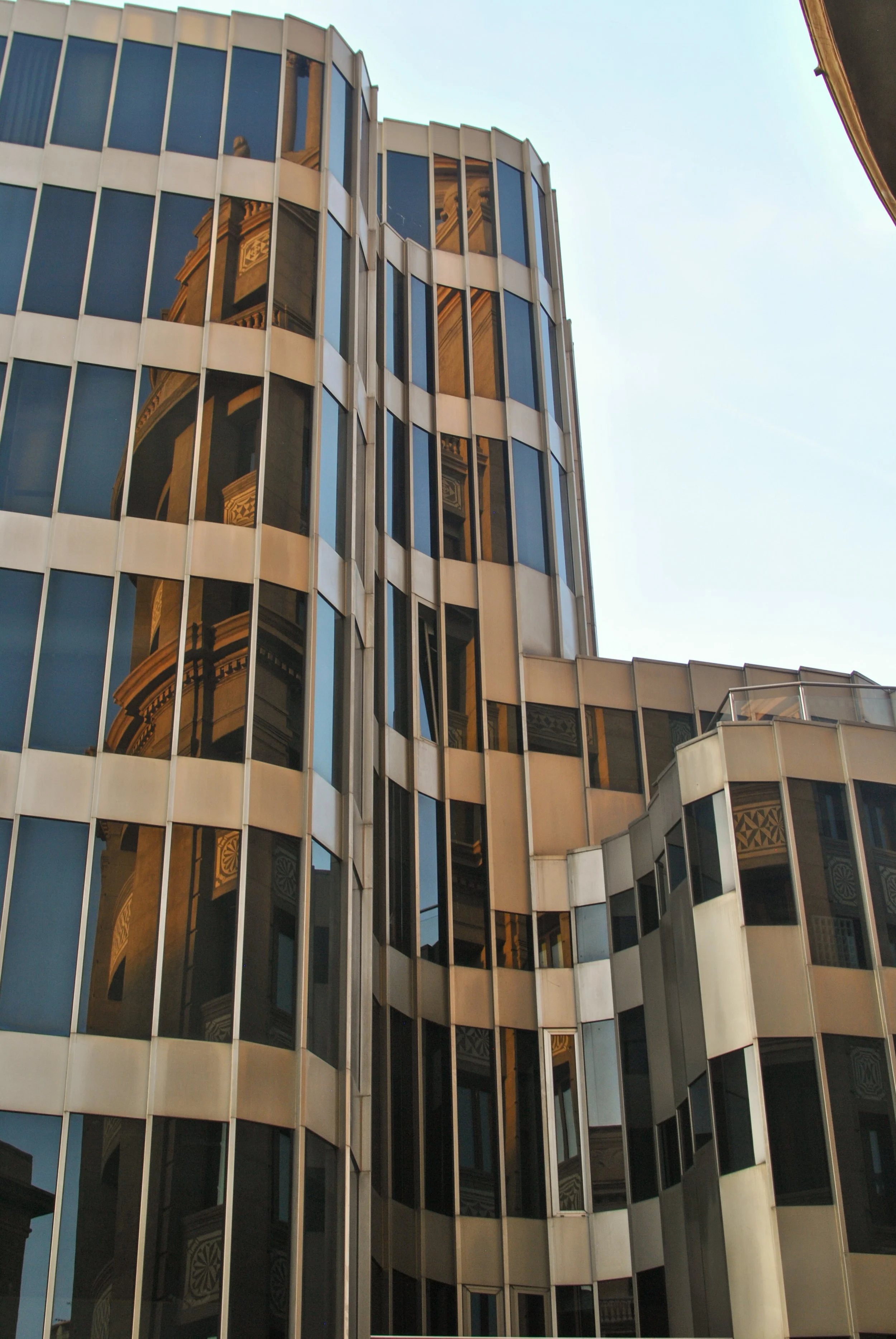 Reflection of an ornate building in the glass windows of a modern, curved office building with vertical metal beams.