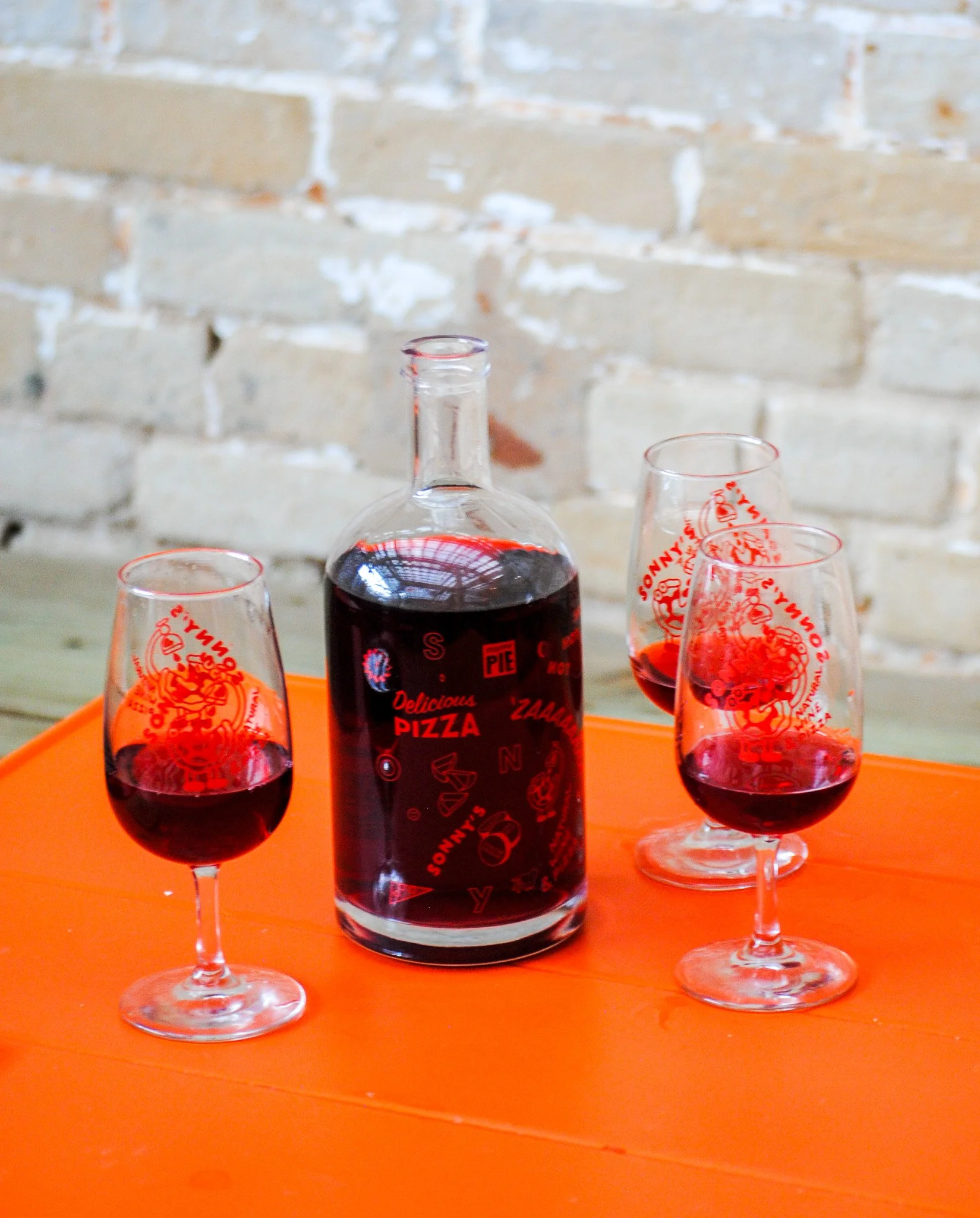 A glass bottle of red beverage and three wine glasses filled with red drink, placed on an orange surface against a white brick wall background.