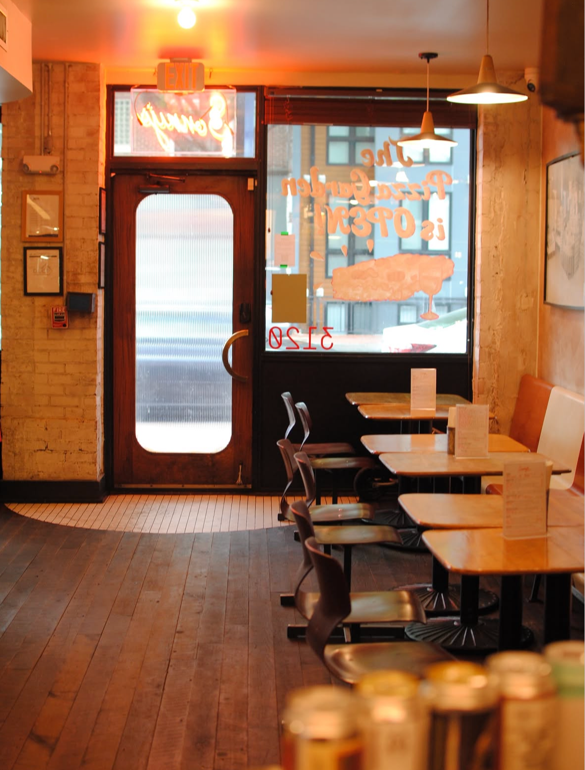 Inside a cozy café with wooden floors and tables, a large window and a wooden door with frosted glass, warm lighting, and a visible neon sign outside.