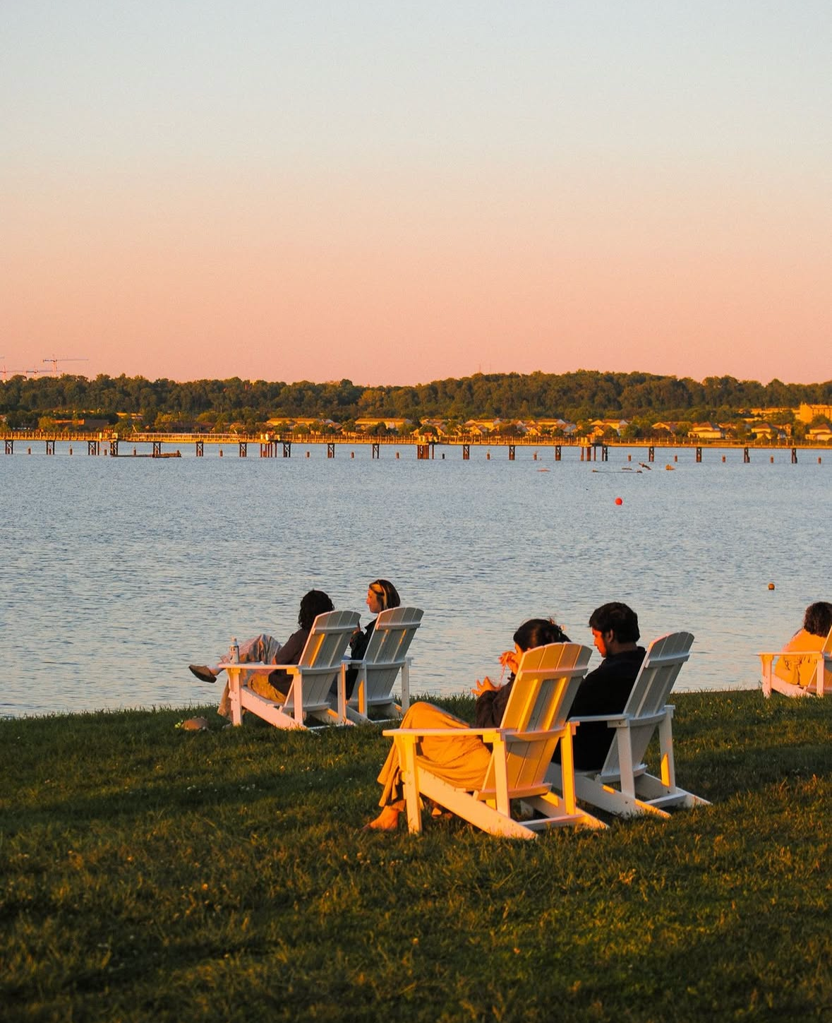 People sitting on white Adirondack chairs by the water during sunset, with a distant shoreline and trees in the background.