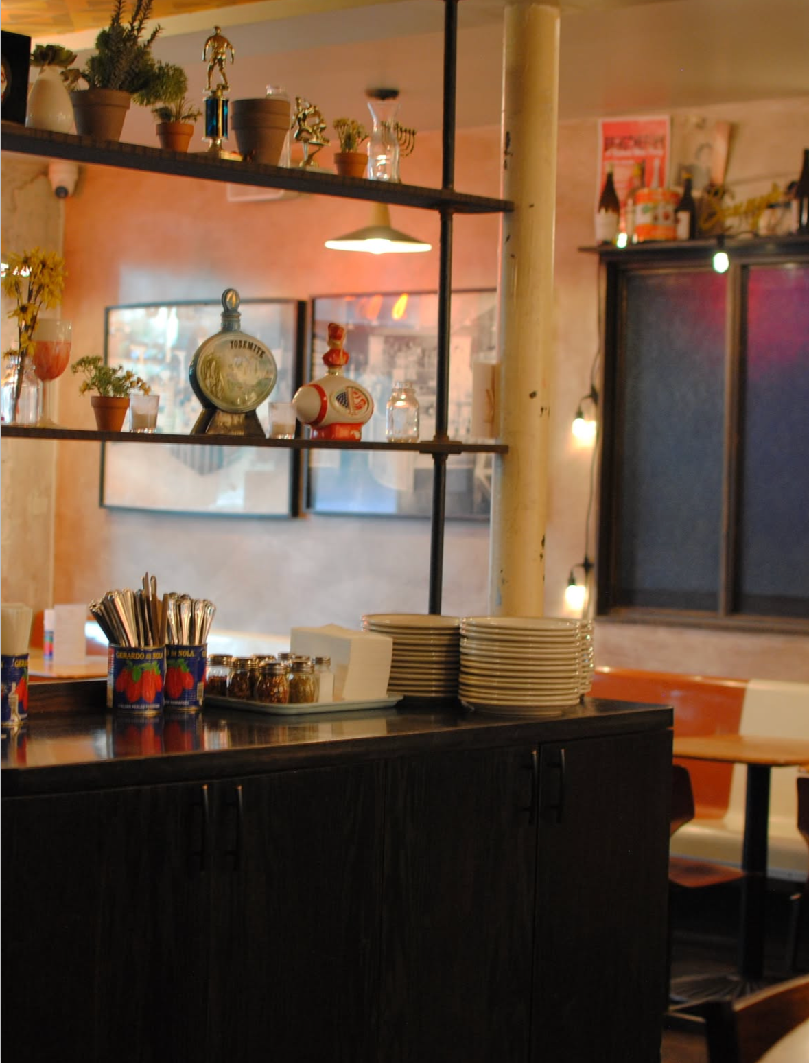 Interior view of a cozy cafe or restaurant, featuring a black countertop with plates, utensils, and jars, with shelves displaying decorative miniatures, plants, and framed artwork in the background.