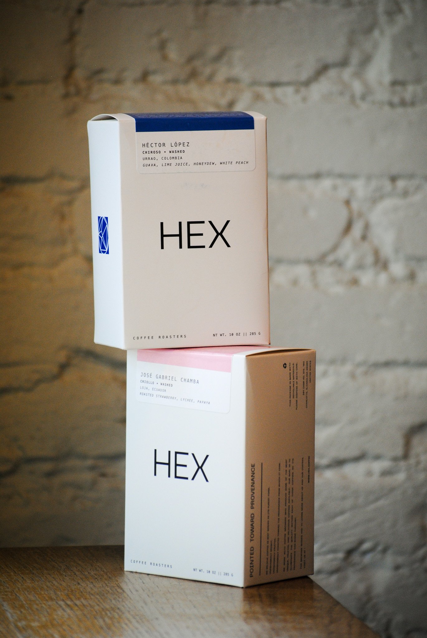 Two rectangular boxes of coffee roasters, one white and one pink, with minimalist design and black text, placed on a wooden table against a stone wall background. The white box is labeled 'HEX' and the pink box is labeled 'José Gabriel Chamba'.